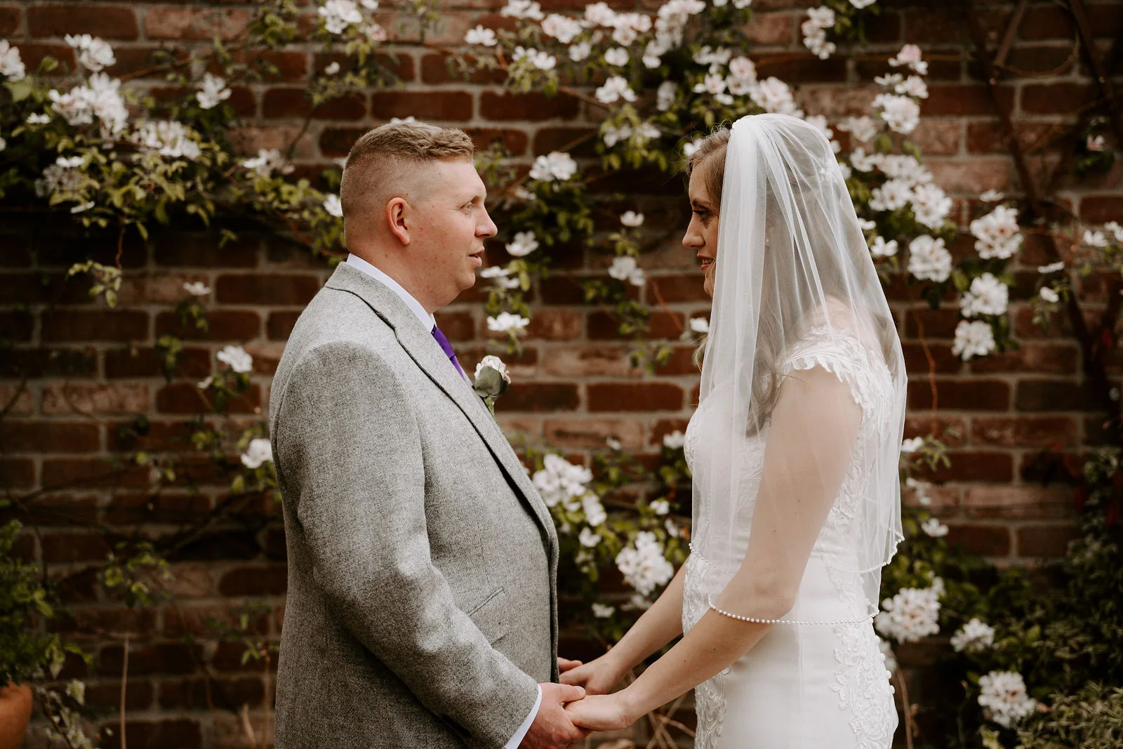 A bride in a veil and a groom in a grey suit stand face to face holding hands in front of a brick wall covered in white roses at Old Thorns