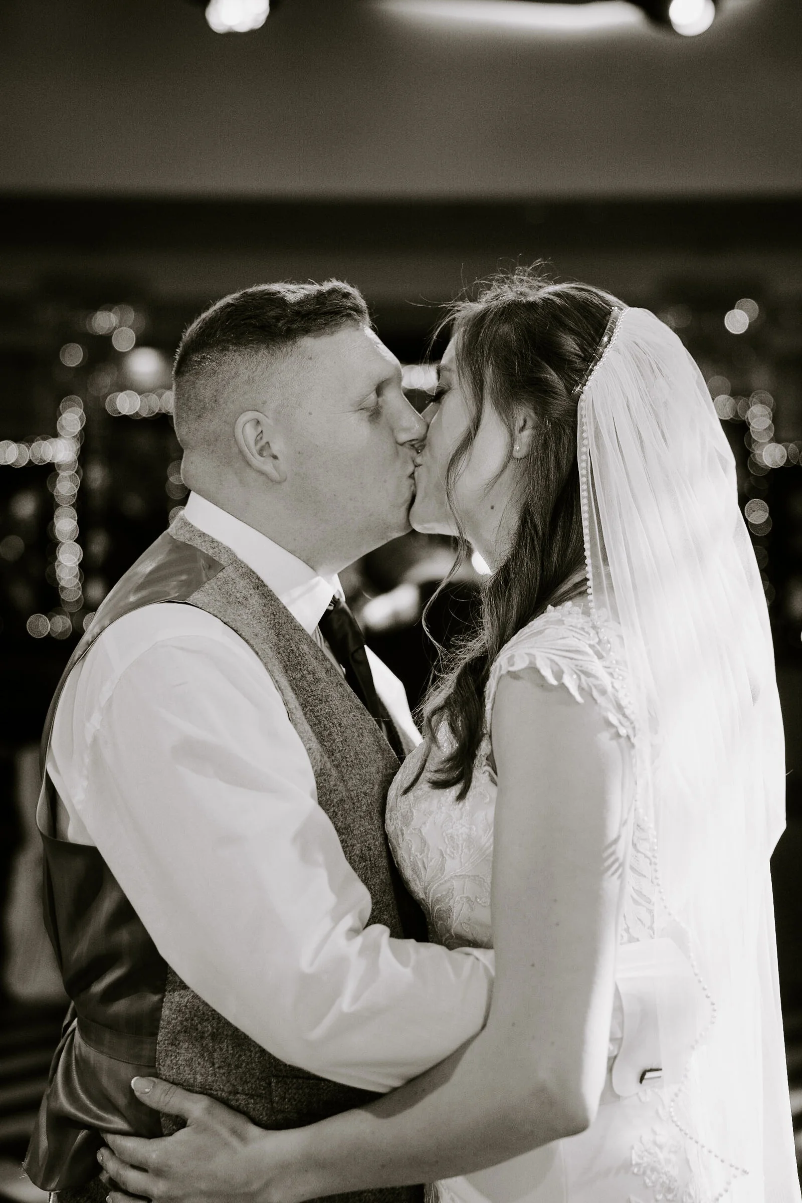 In black and white, a groom in a waistcoat and a bride in a veil kiss with their arms around each other at Old Thorns