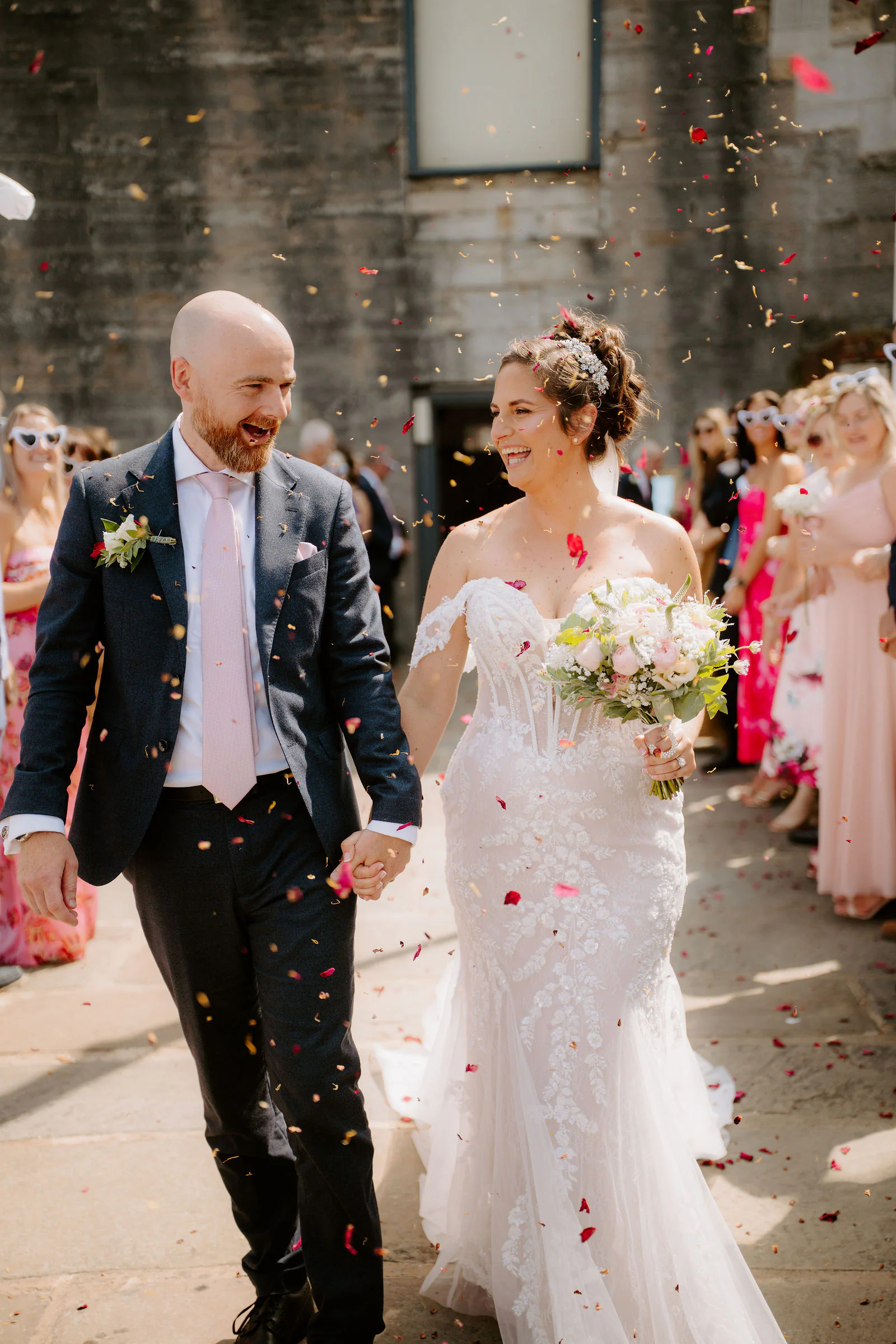 Claire and James walk hand in hand through a shower of confetti between lines of cheering guests outside a stone building