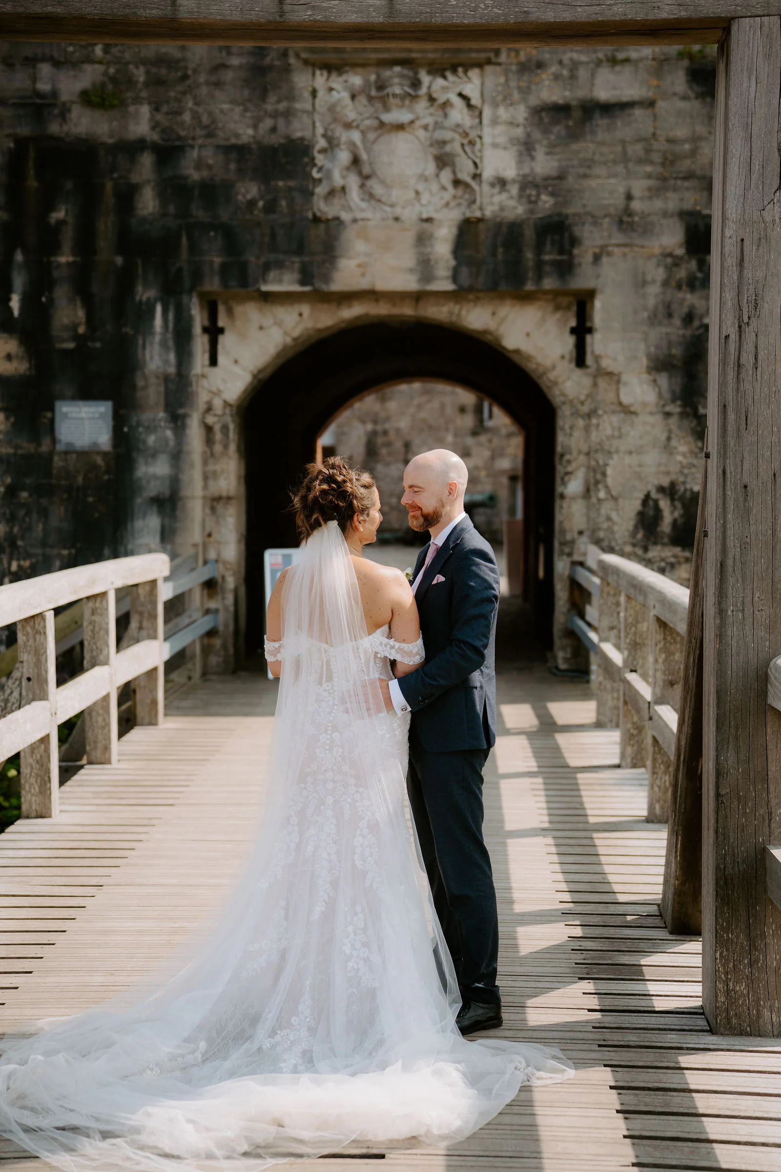 Claire in a lace gown and James in a navy suit face each other on a wooden bridge beneath a stone archway, Claire's veil trailing behind her