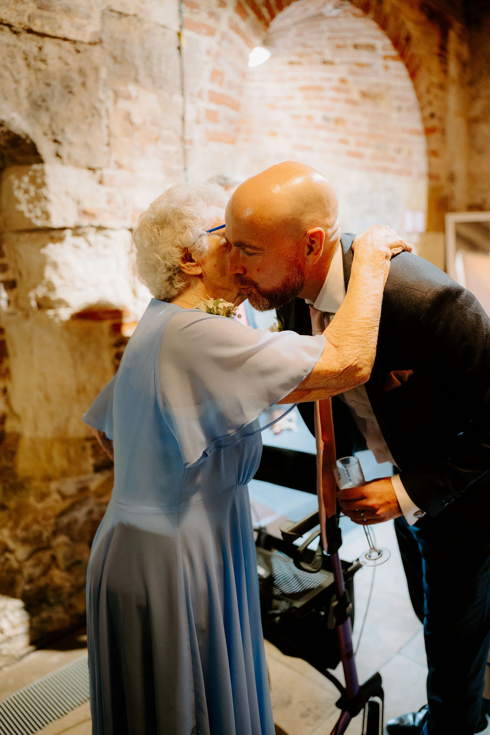 James bends to kiss an older relative on the cheek inside a stone-walled room with brick arches, warm light falling across them