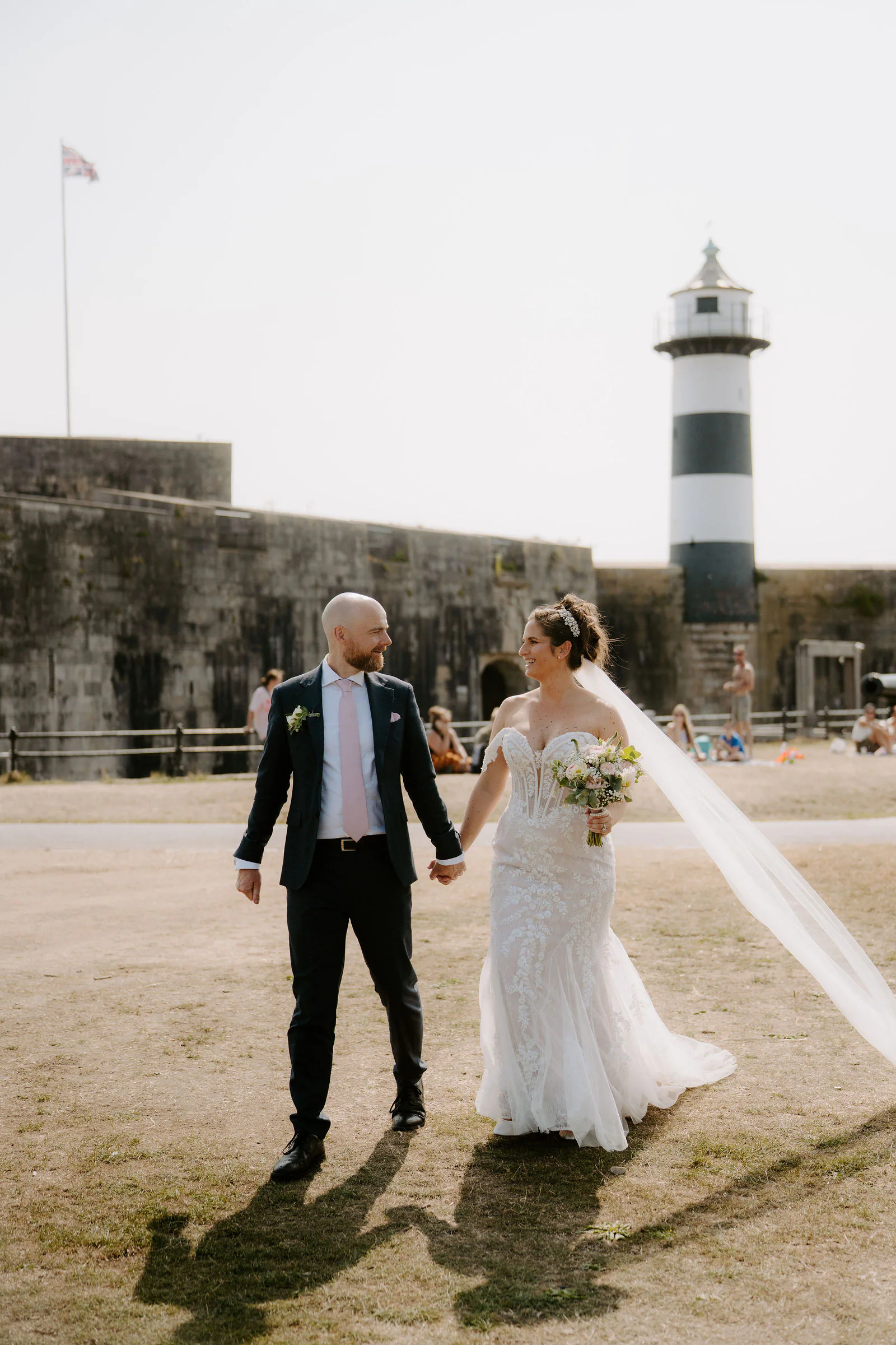 Claire in a lace gown with a long veil and James in a navy suit walk hand in hand across a sunlit lawn in front of a stone fort wall and a black and white striped lighthouse