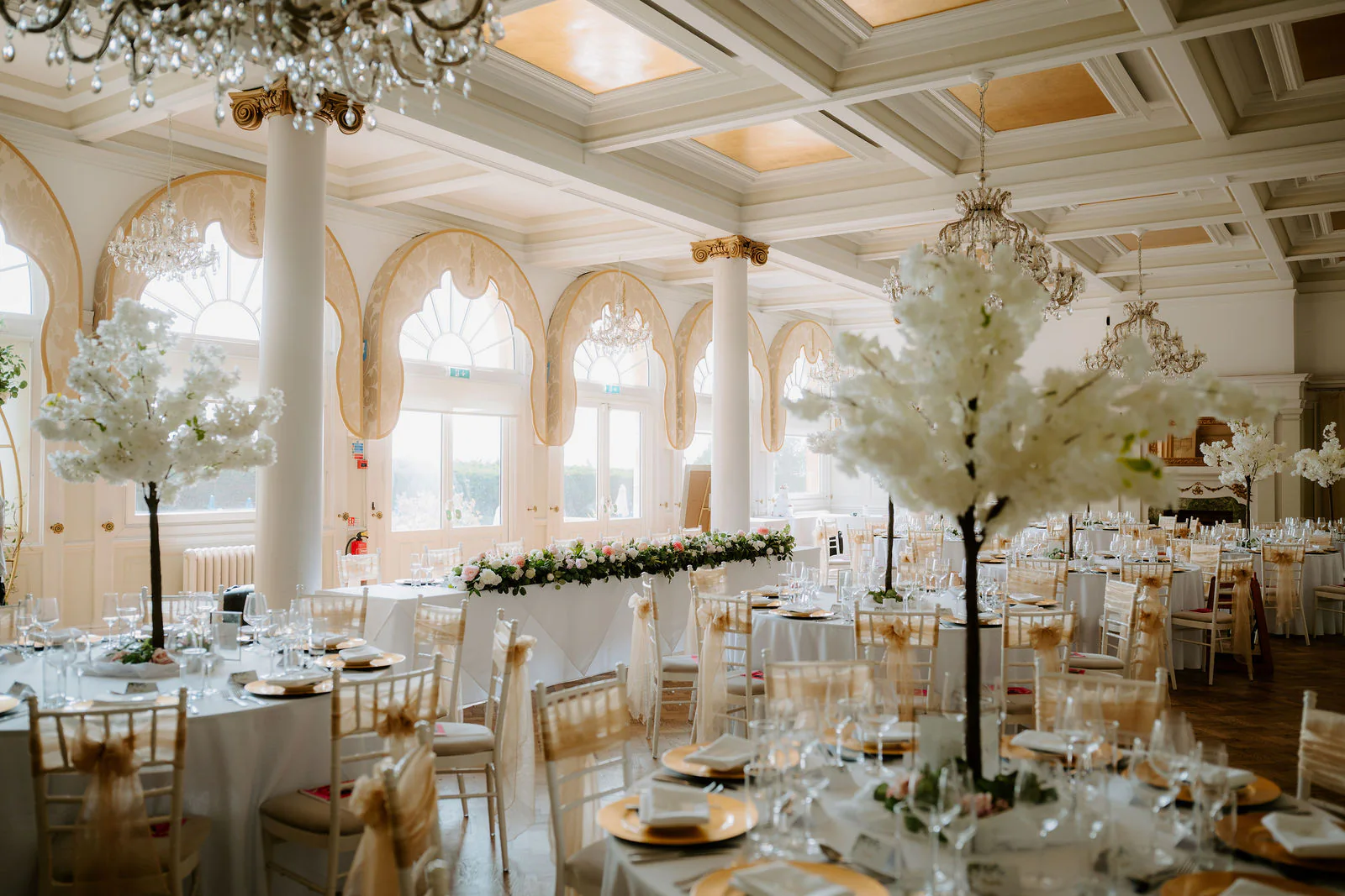 A reception room laid with round tables, chandeliers and white blossom trees beneath tall arched windows