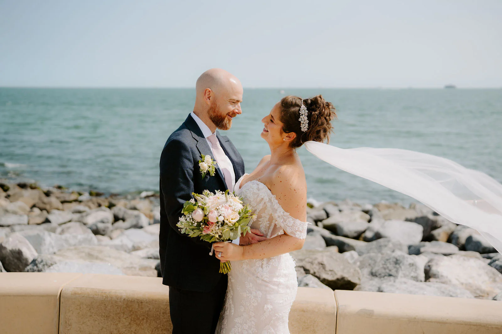 Claire and James look at each other on the seafront as the wind lifts her veil, with rocks and the sea behind them