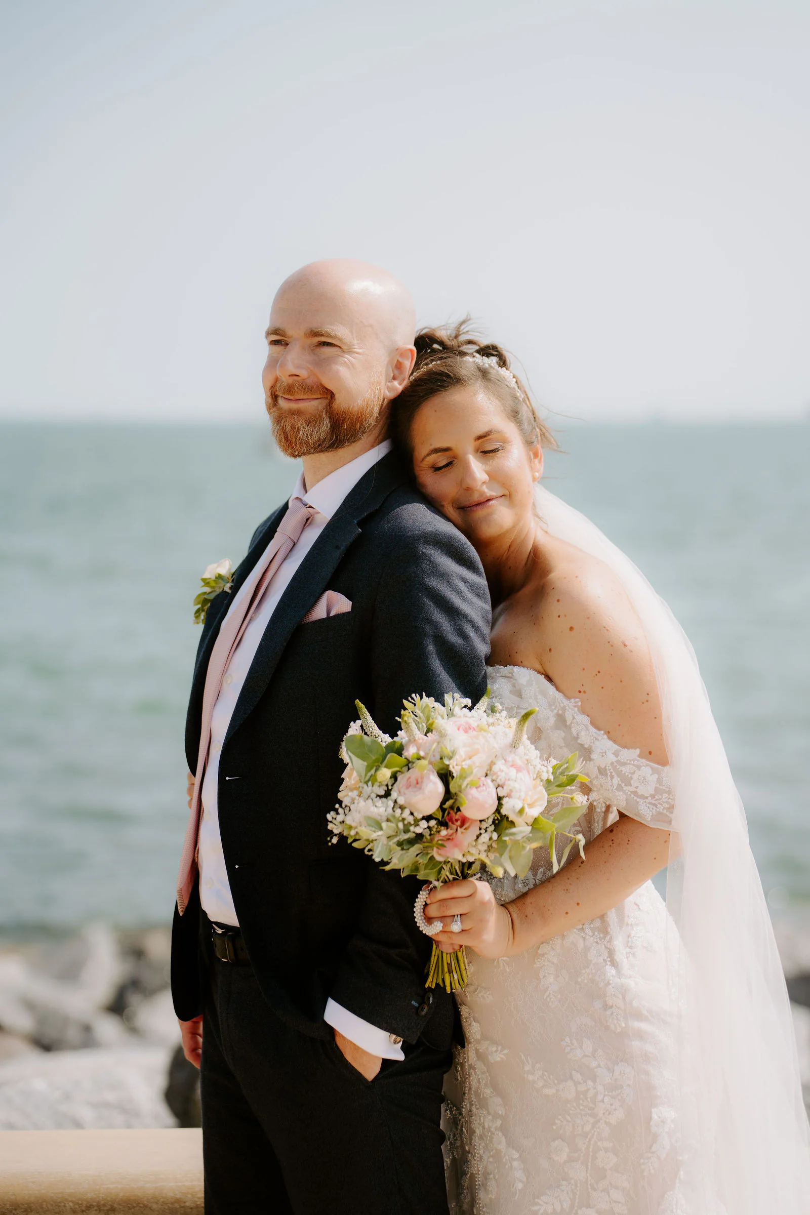 Claire rests her head against James's shoulder with her eyes closed, a bouquet of pink and white flowers in her hand, with the sea behind them