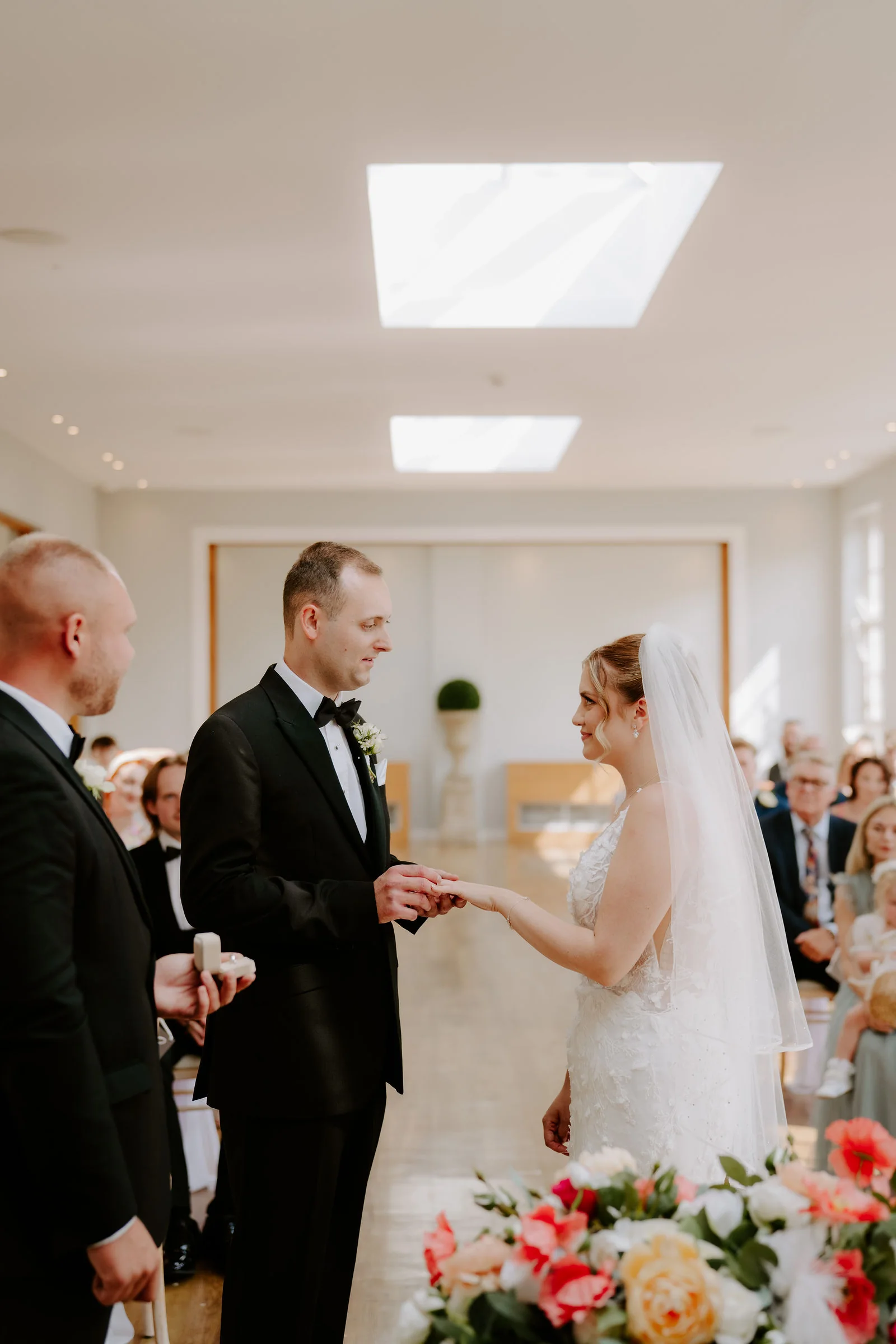 A groom in black tie puts a ring on the finger of his bride, who wears a veil, in a bright room with a best man beside the groom and a bouquet of coral and white flowers in the foreground