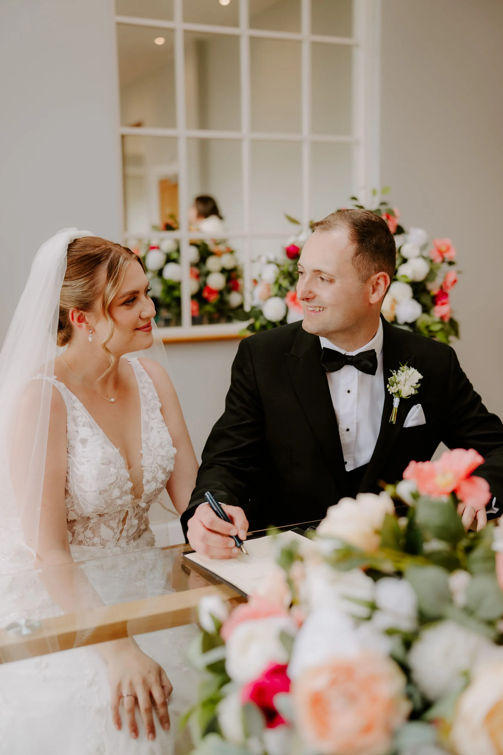 A groom in black tie signs the register beside his bride, who wears a veil and watches him, with a bouquet of coral, white and pink flowers in the foreground and tall windows behind