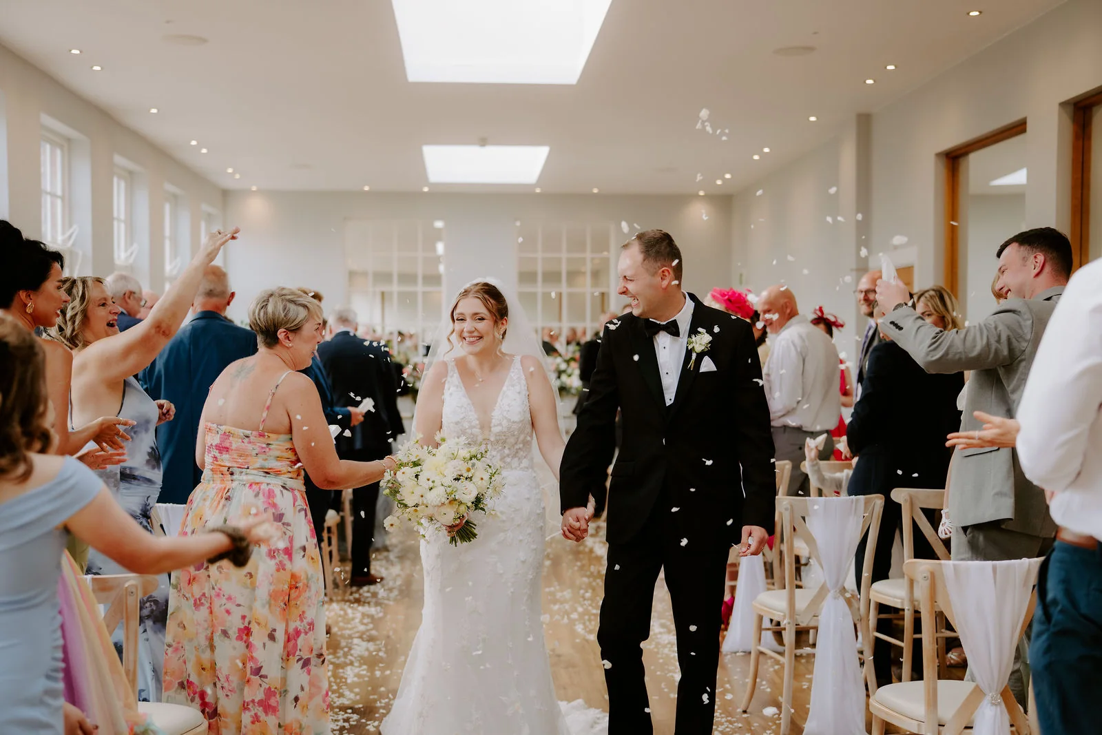 A bride in a veil holding a bouquet of white flowers and a groom in black tie walk hand in hand up an aisle between guests throwing petals, in a bright room with skylights and tall windows