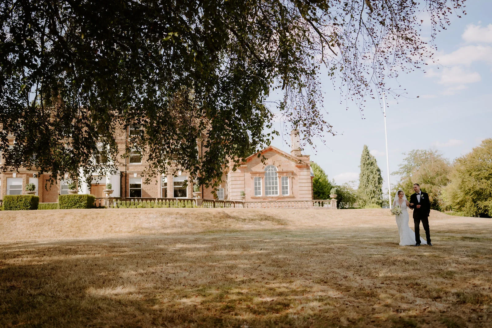 A distant view of a bride and groom standing on a dry lawn beneath a large beech tree, with a red-brick manor house and a flagpole behind them under a blue sky