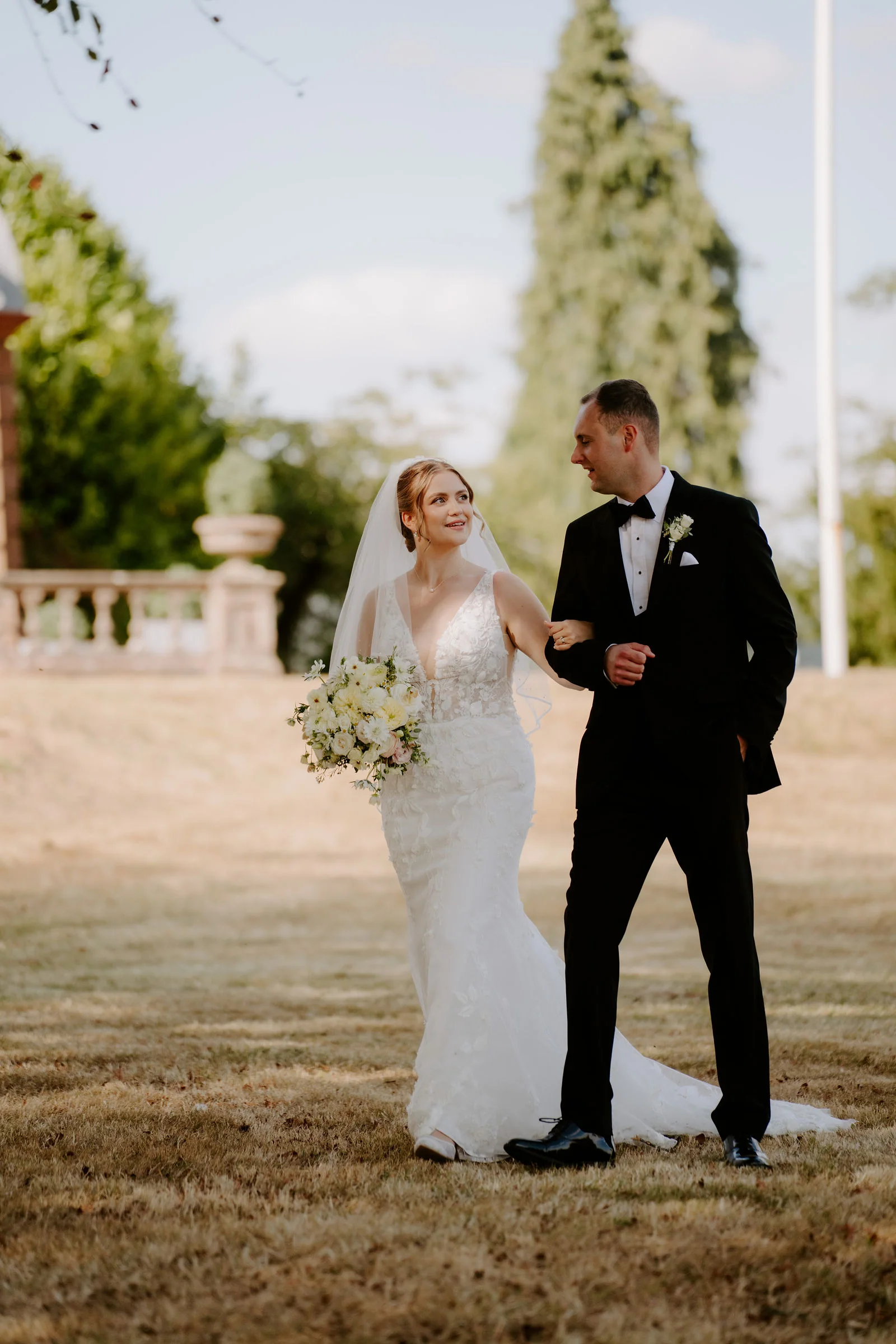 A bride in a veil holding a bouquet of white flowers walks arm in arm with a groom in black tie on a dry lawn, with a stone balustrade, a tall cypress and a flagpole behind them