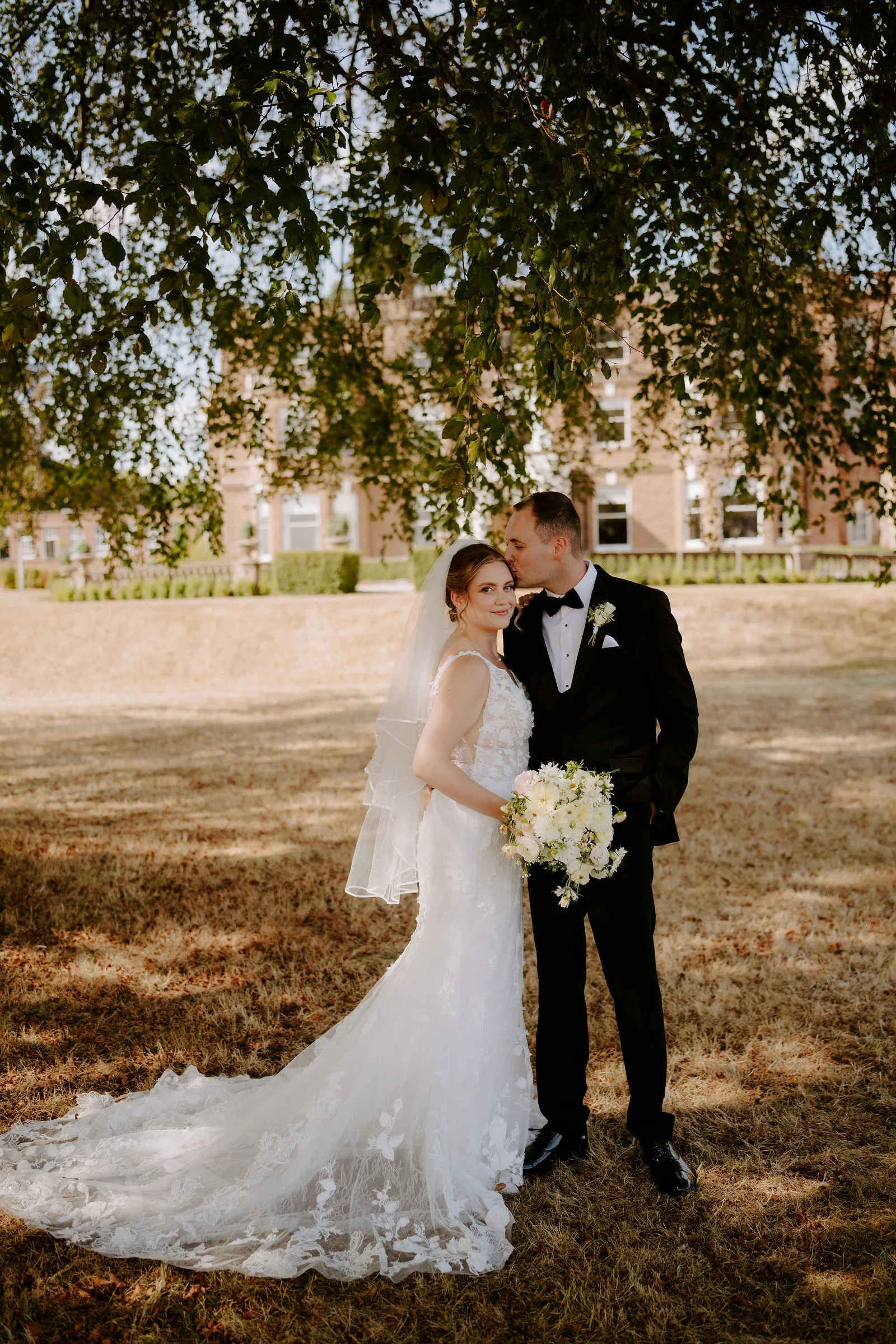 A groom in black tie kisses the forehead of his bride, who wears a veil and holds a bouquet of white flowers, beneath the branches of a large beech tree with a red-brick manor house blurred behind them