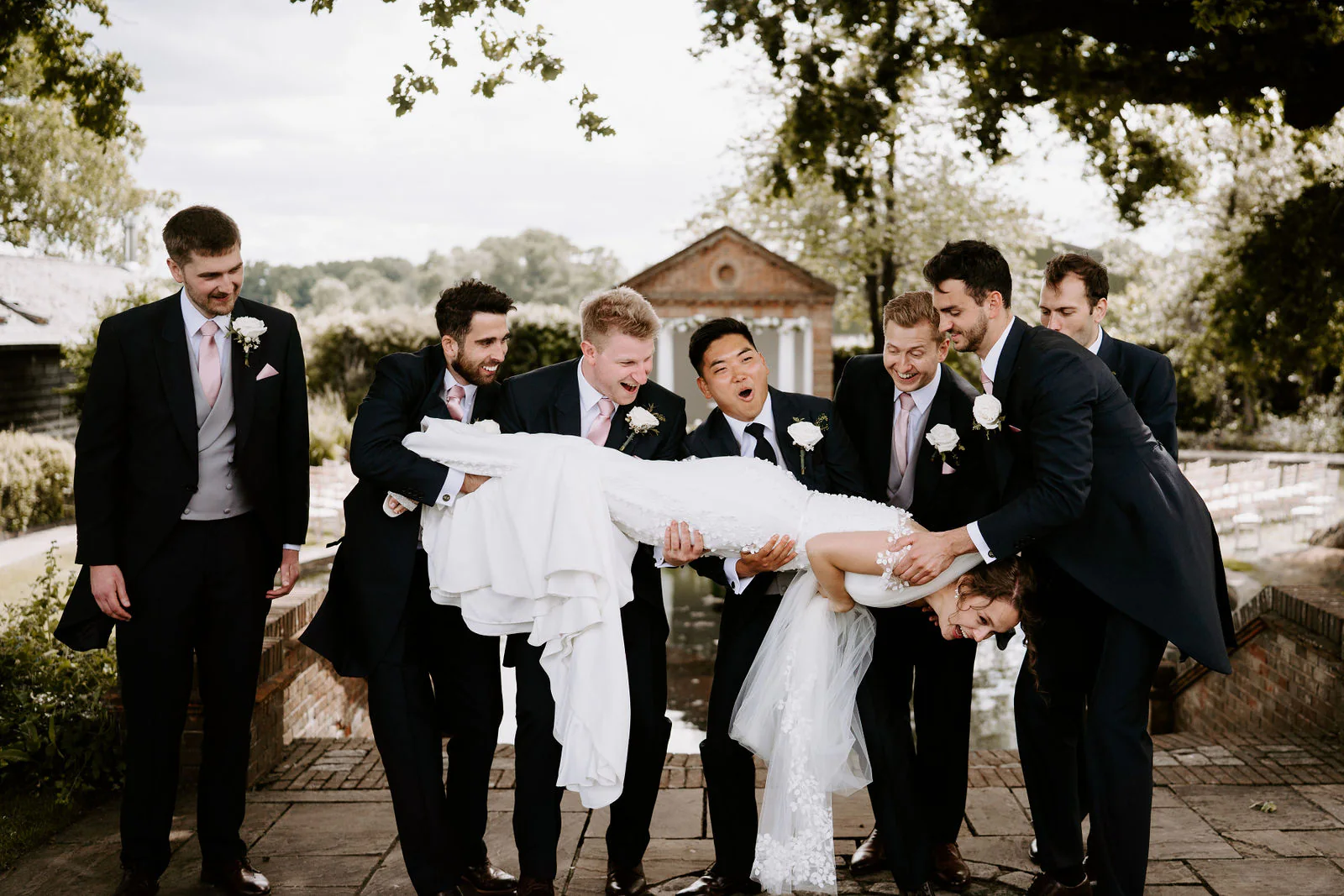 Tae and his groomsmen in dark suits carry Becky horizontally in her wedding dress beside the pavilion while everyone laughs