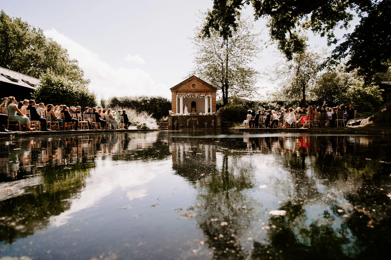 Wedding guests gathered around the still pond and pavilion at Micklefield Hall, which are reflected in the water