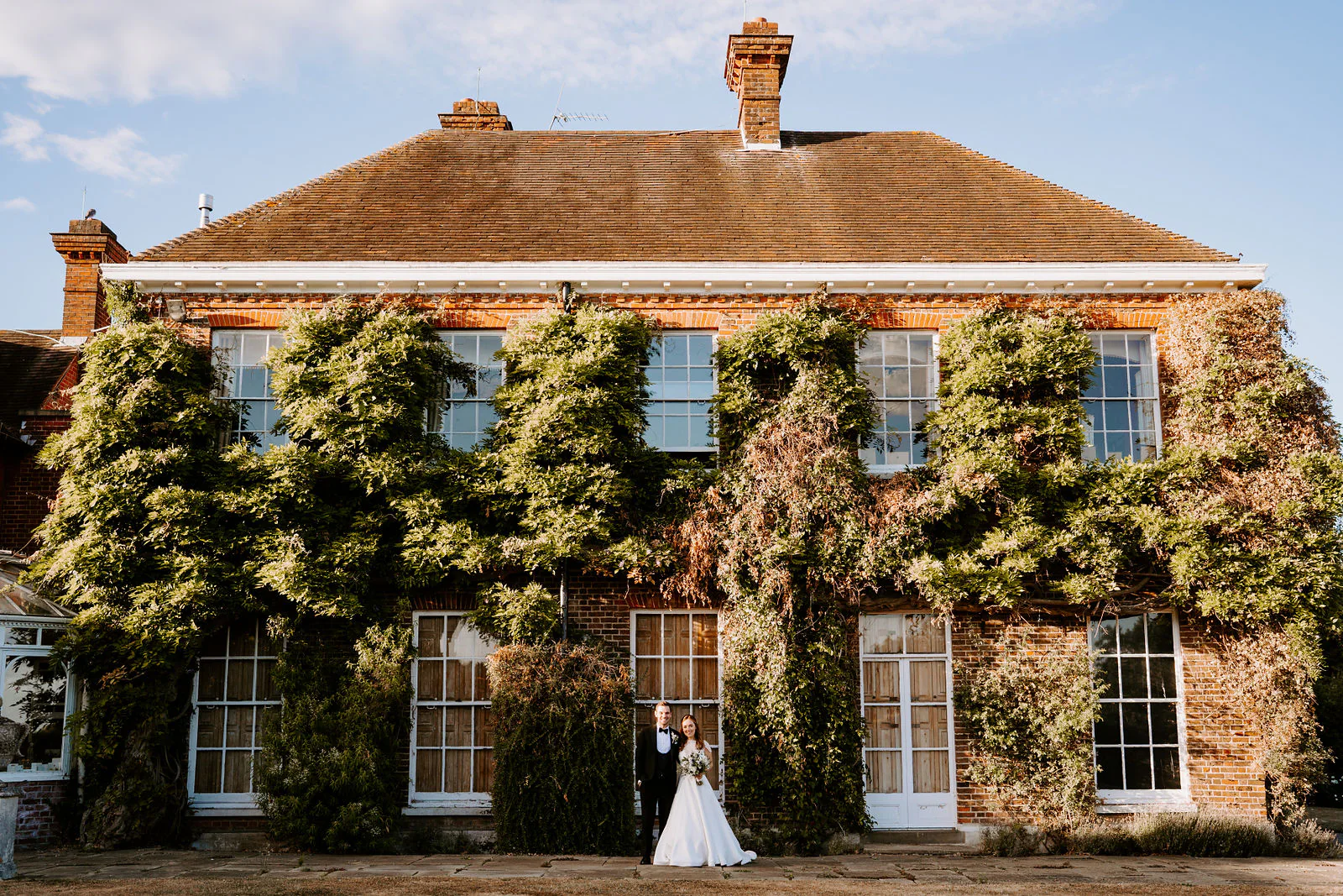 Charlotte and Matt stand small in front of the ivy-covered red-brick house at Micklefield Hall under a blue sky