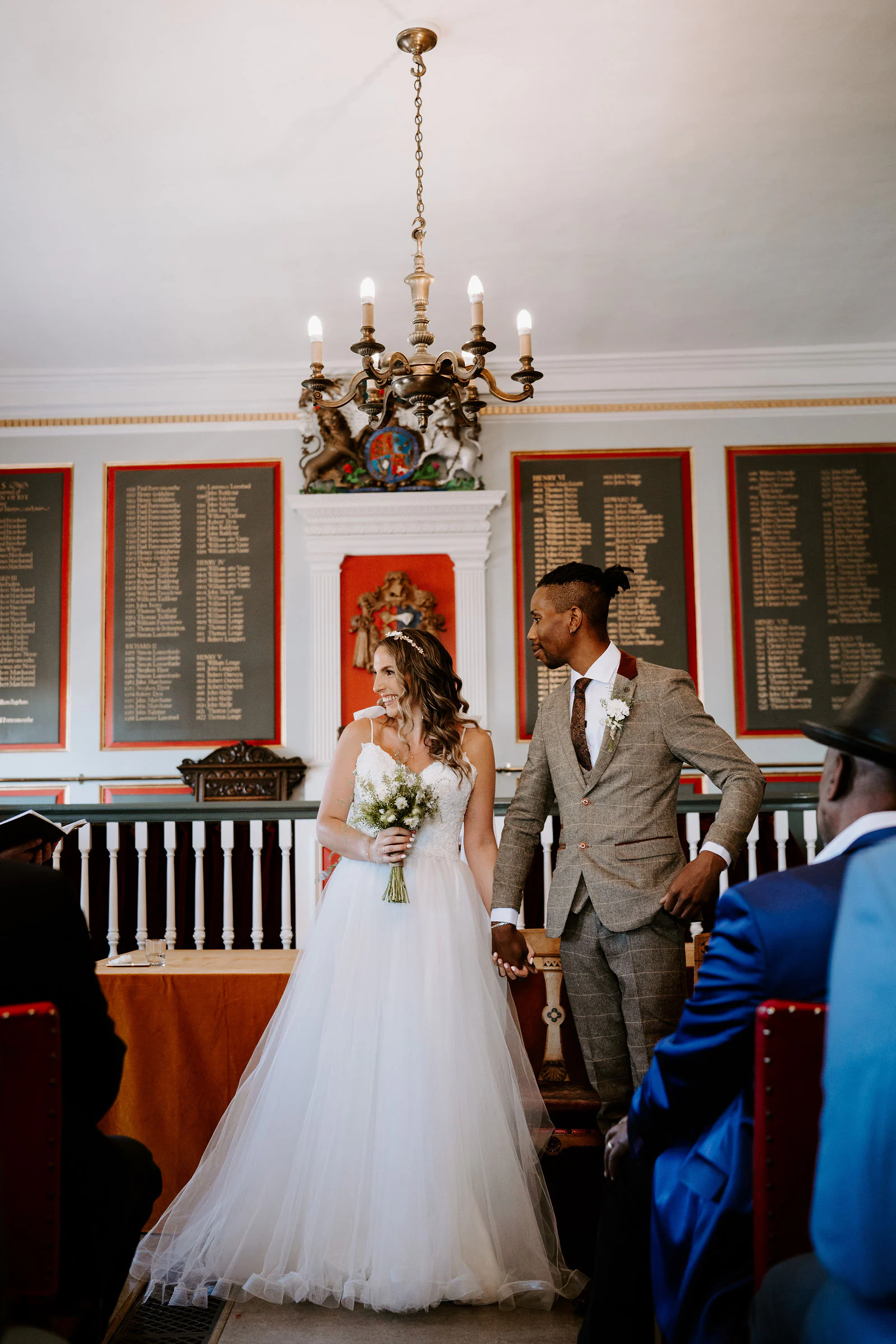 A bride and groom stand in a panelled room with a chandelier and boards of names behind them during their ceremony in Rye