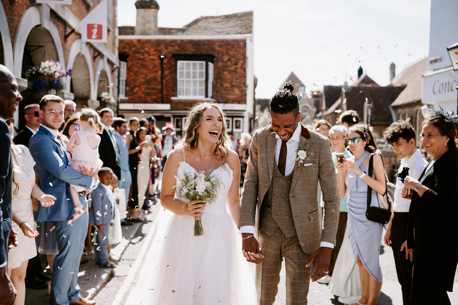 A bride and groom laugh as guests gather around them on a busy Rye street
