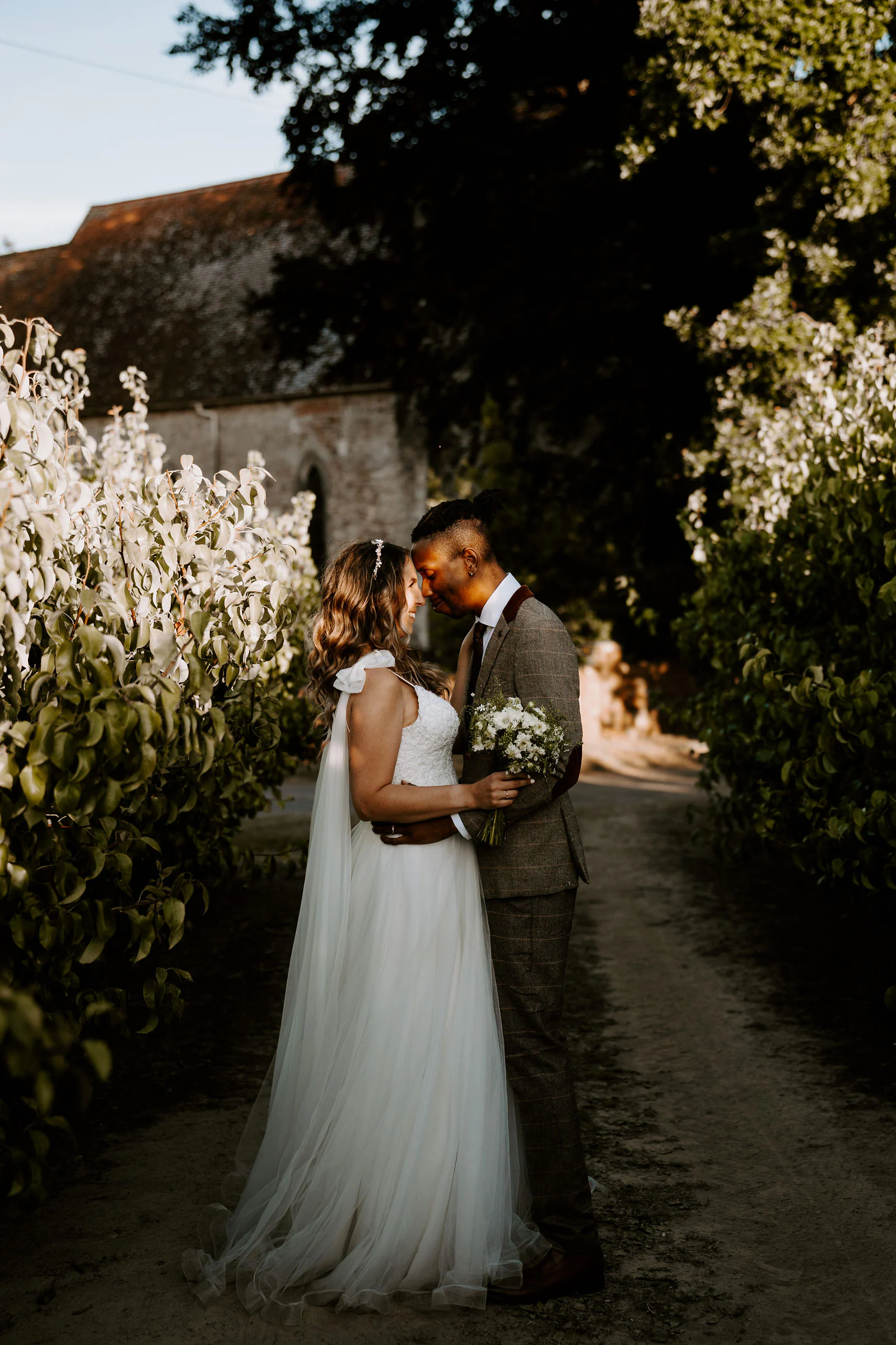 A couple embrace on a gravel path beside hedges in warm evening light in East Sussex