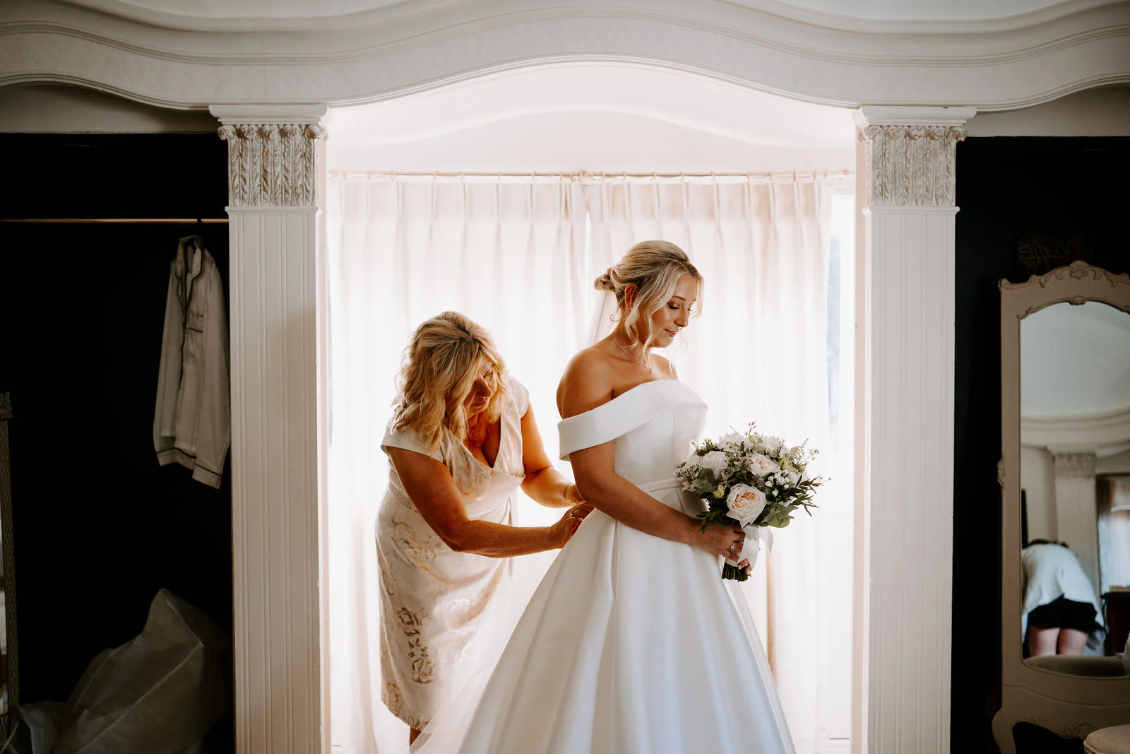 A bridesmaid adjusts the bride's dress beside a tall window at Hayne House in Hythe