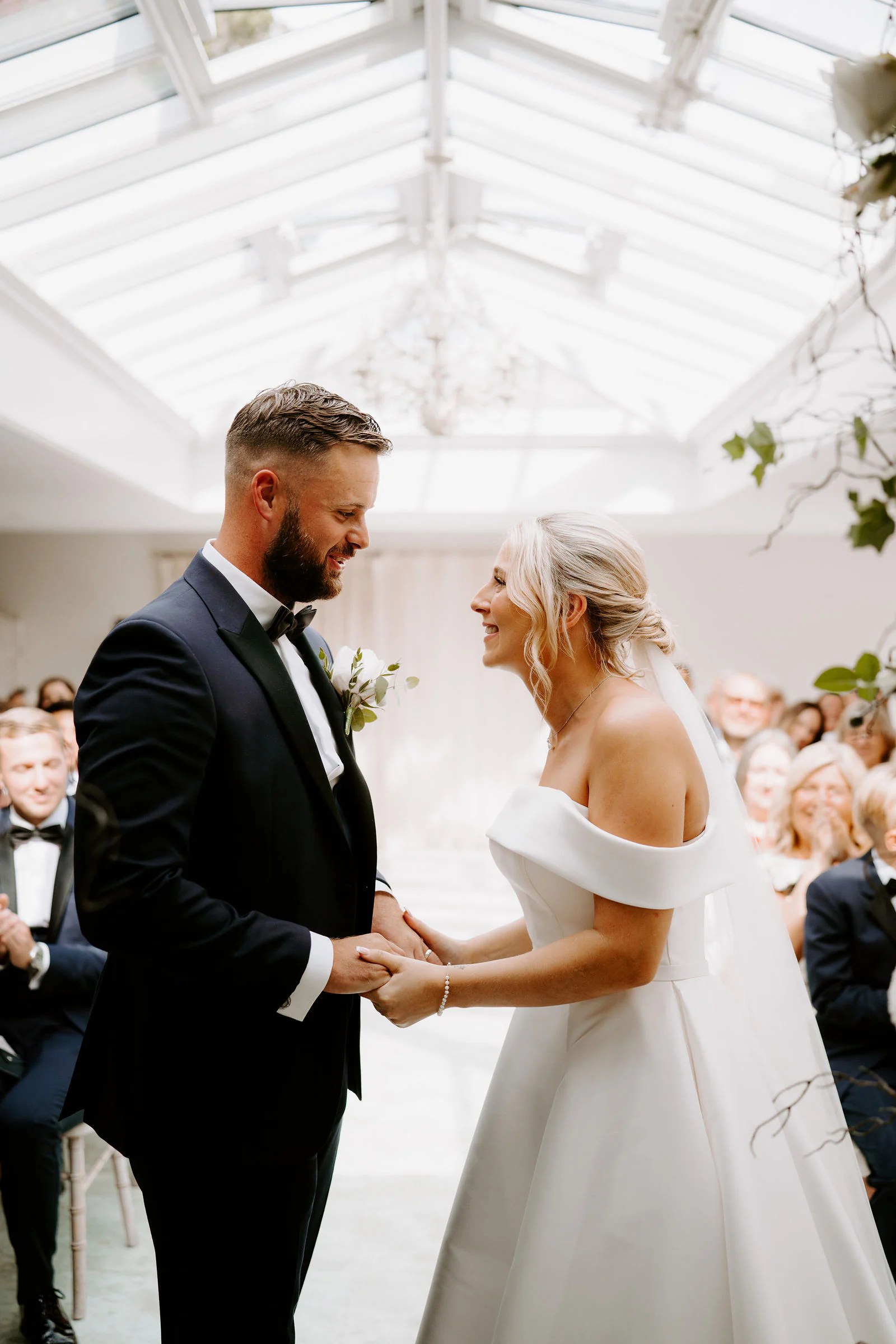A bride and groom hold hands and smile at each other beneath a glass roof as guests look on at Hayne House in Hythe