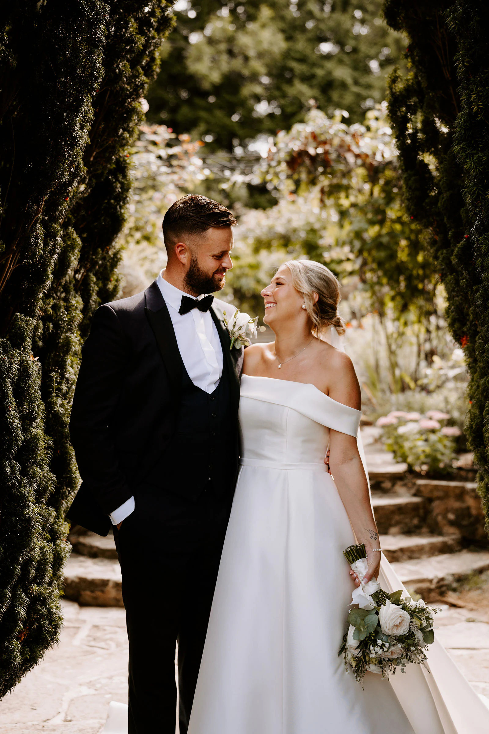 A groom in a black bow tie and a bride in an off-the-shoulder gown stand together beside stone steps in the garden at Hayne House in Hythe