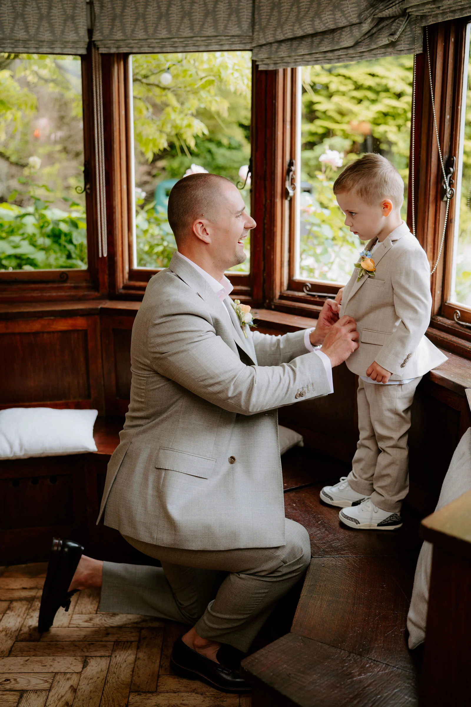 A groom in a light suit kneels in a wooden bay window to talk with a small boy in a matching suit at Hayne House near Hythe