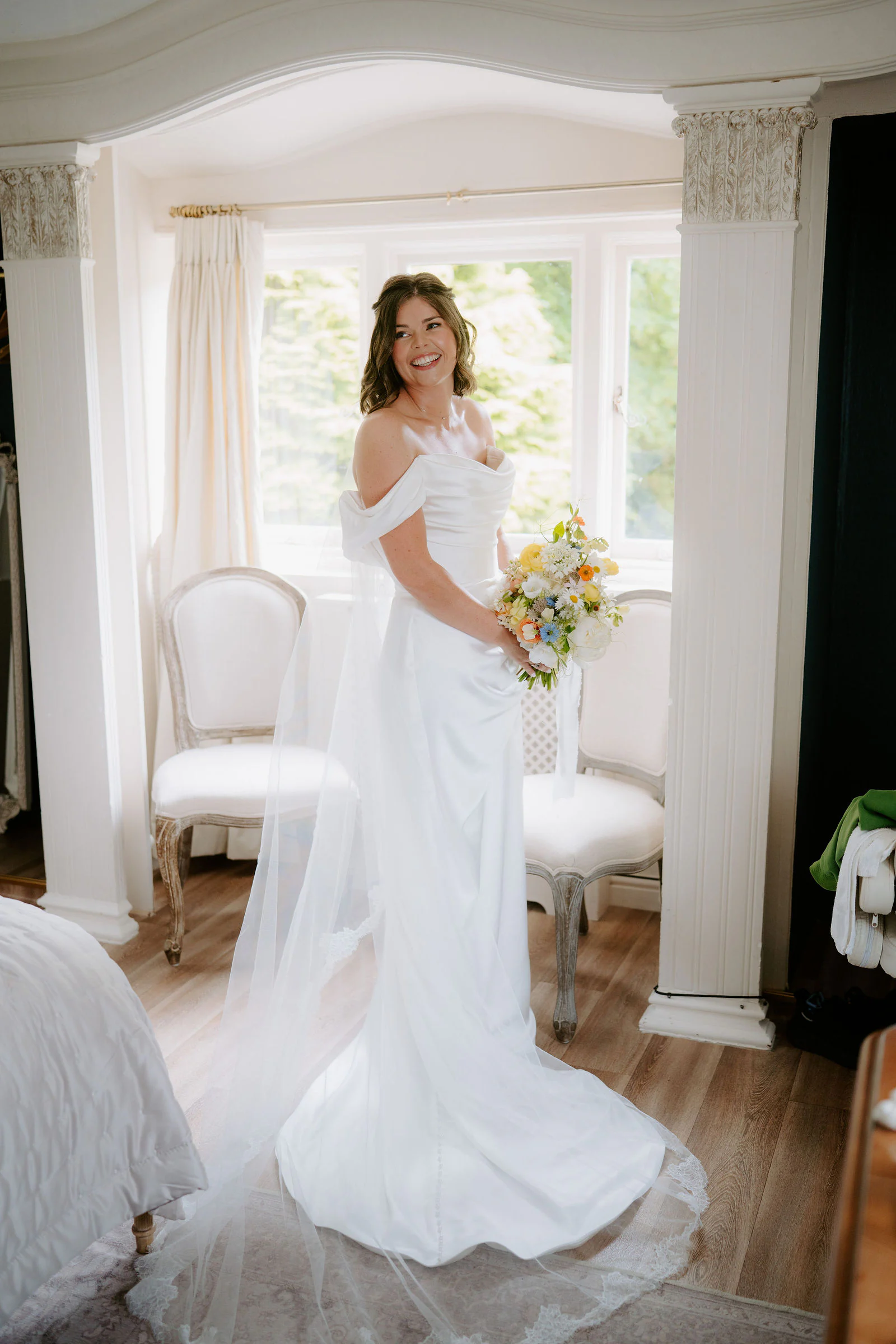 A bride in a satin wedding dress holds her bouquet in a bright room with a bay window at Hayne House near Hythe