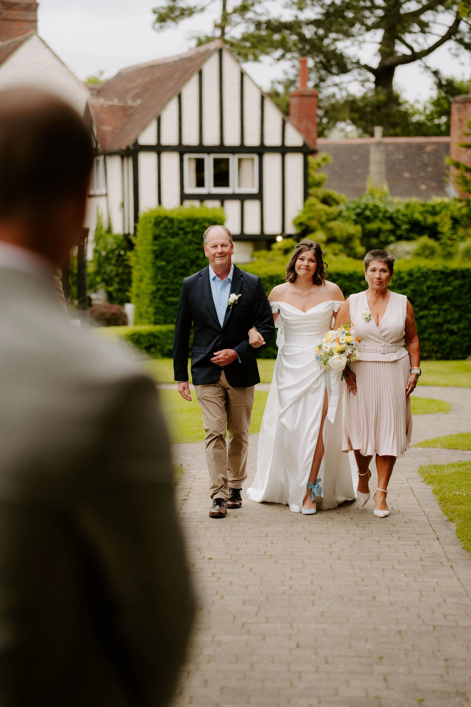 A bride walks along a garden path beside two guests with a half-timbered house behind at Hayne House near Hythe