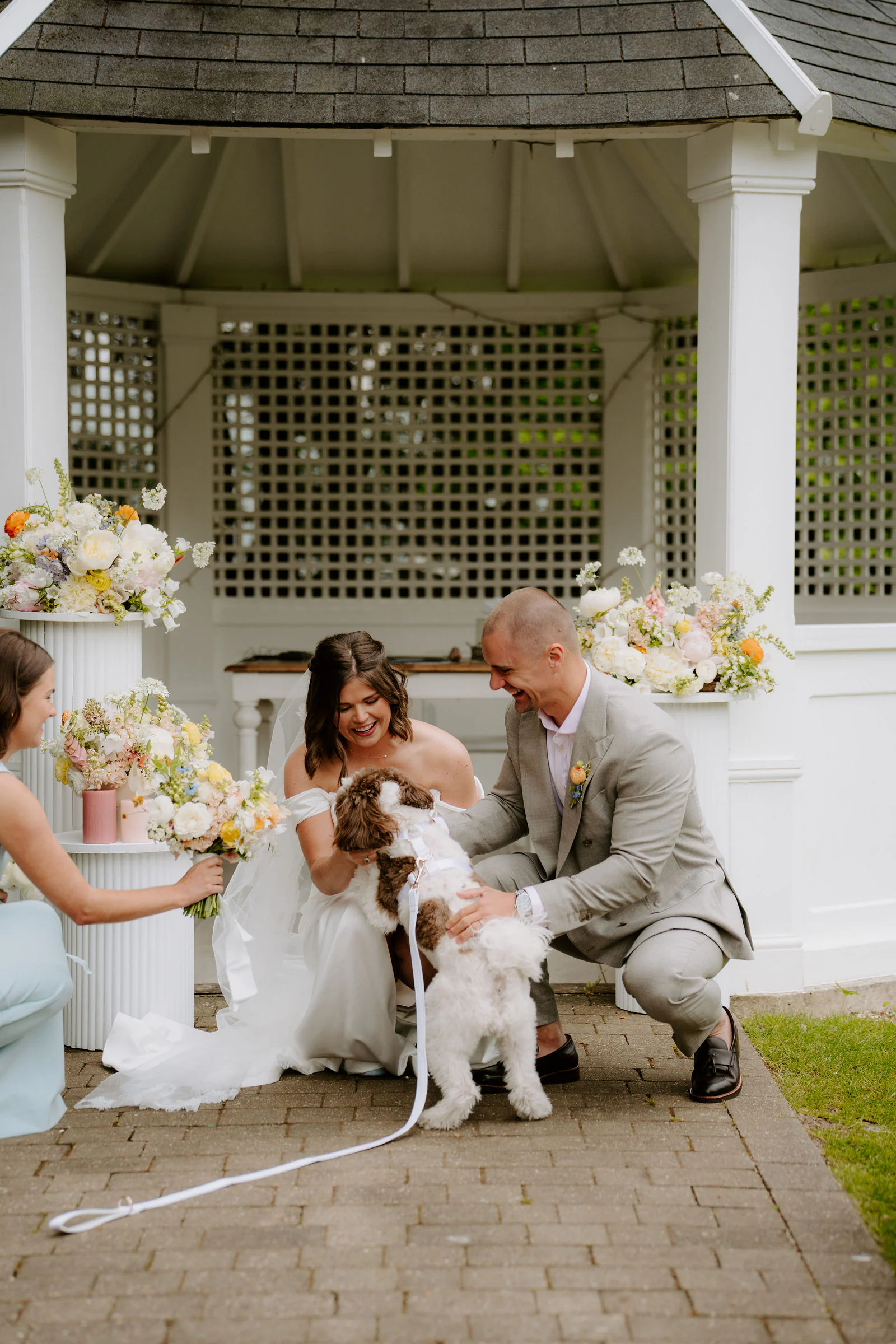 A bride and groom crouch to greet a small dog beside a white garden gazebo at Hayne House near Hythe