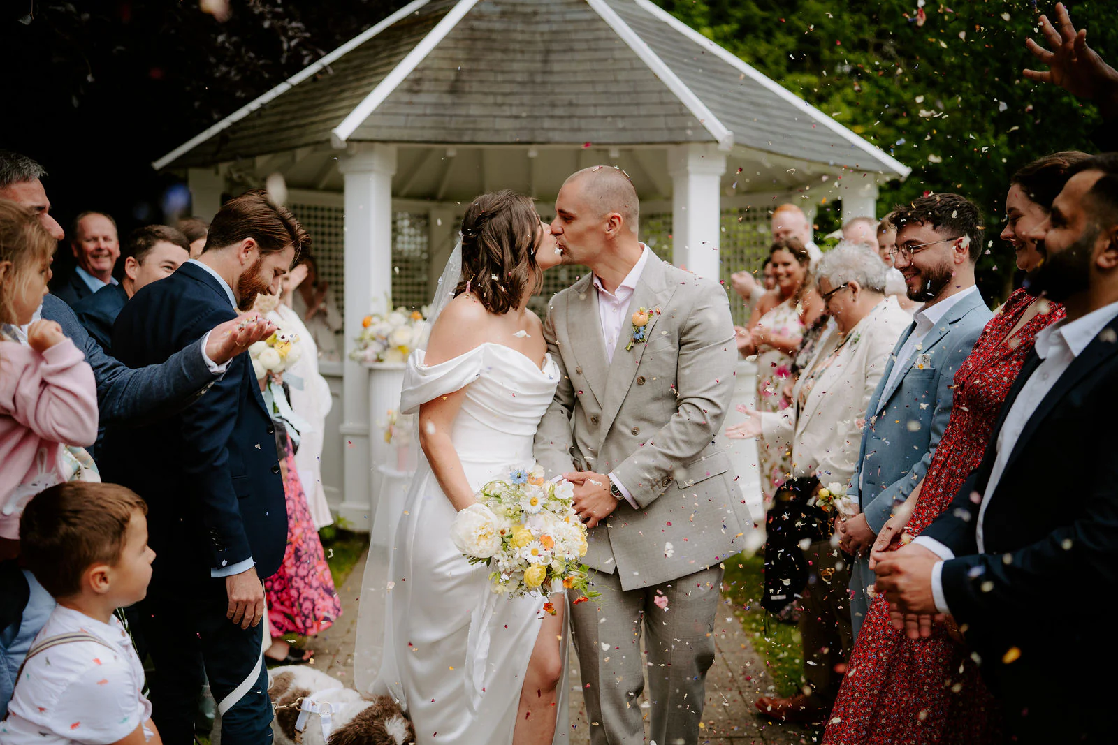 A bride and groom in light clothes are showered with confetti by guests beside a white gazebo at Hayne House near Hythe