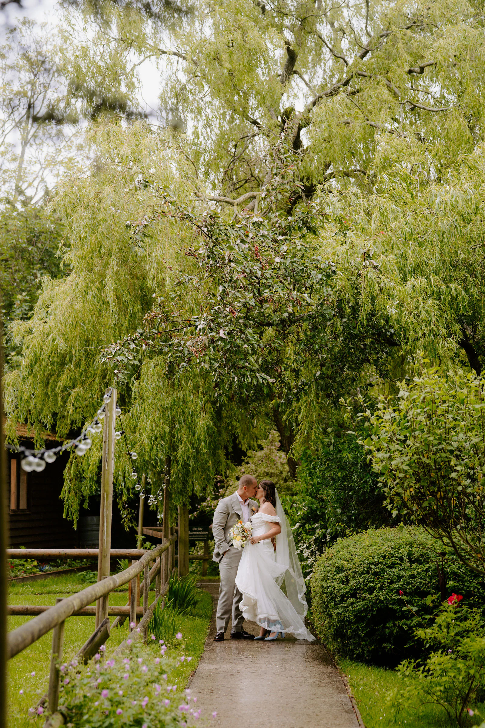 A groom in a stone-coloured suit and a bride in an off-the-shoulder gown and long veil kiss on a garden path beneath a weeping willow with festoon lights beside a fence at Hayne House