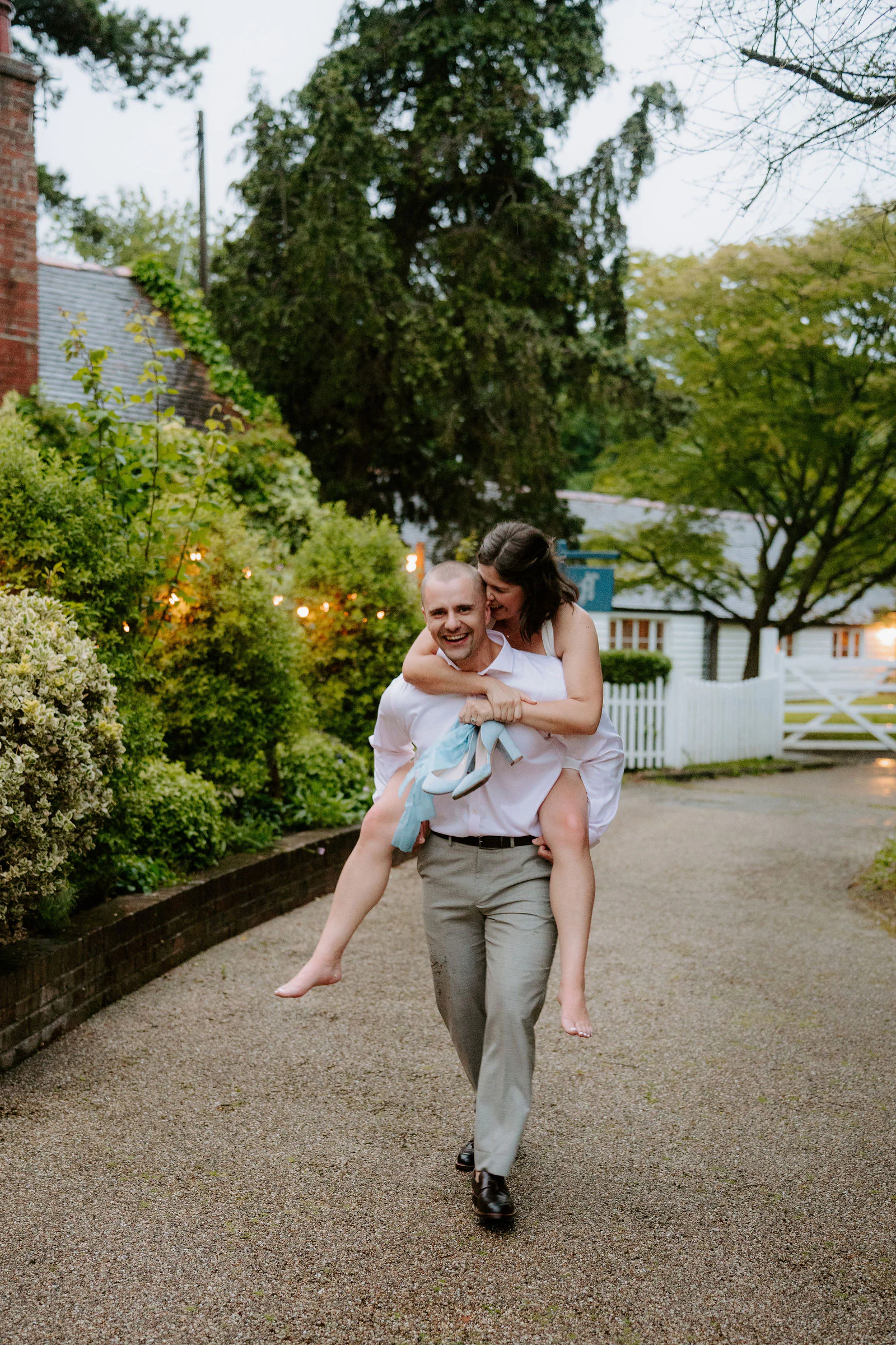 A groom carries his bride piggyback along a lamplit drive in the evening at Hayne House near Hythe