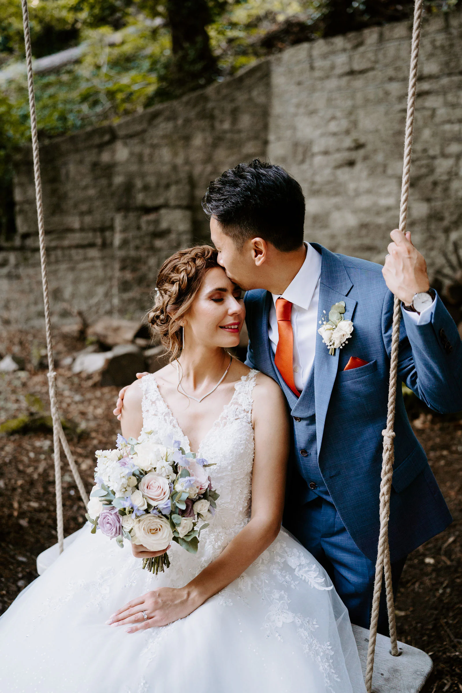 A bride in a lace dress and a groom in a blue suit share a kiss on a rope swing beside an old stone wall at The Orangery in Maidstone