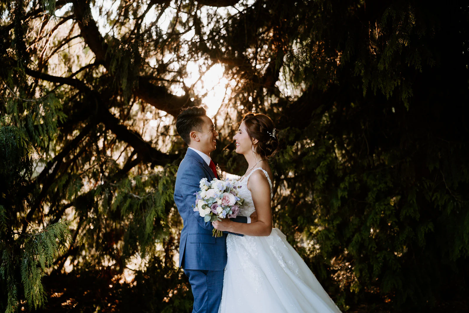A bride and groom embrace in low evening sun beneath tall trees at The Orangery in Maidstone