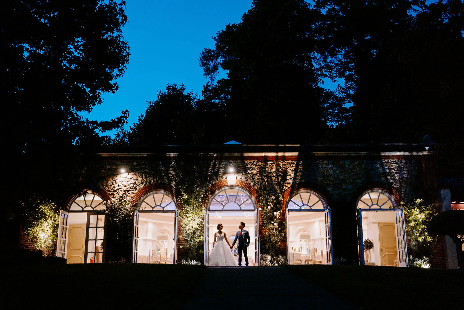 A couple stand side by side at dusk beneath the lit arched windows of The Orangery in Maidstone