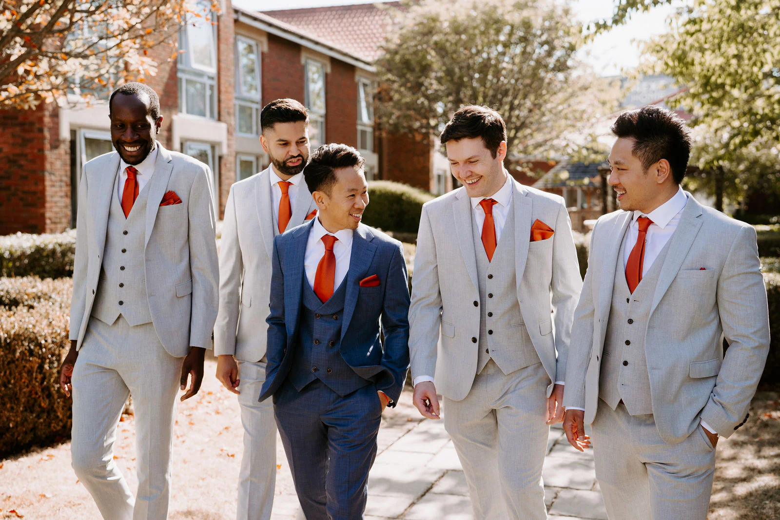 Five groomsmen in light grey suits with red details walk together along a gravel path at The Orangery in Maidstone