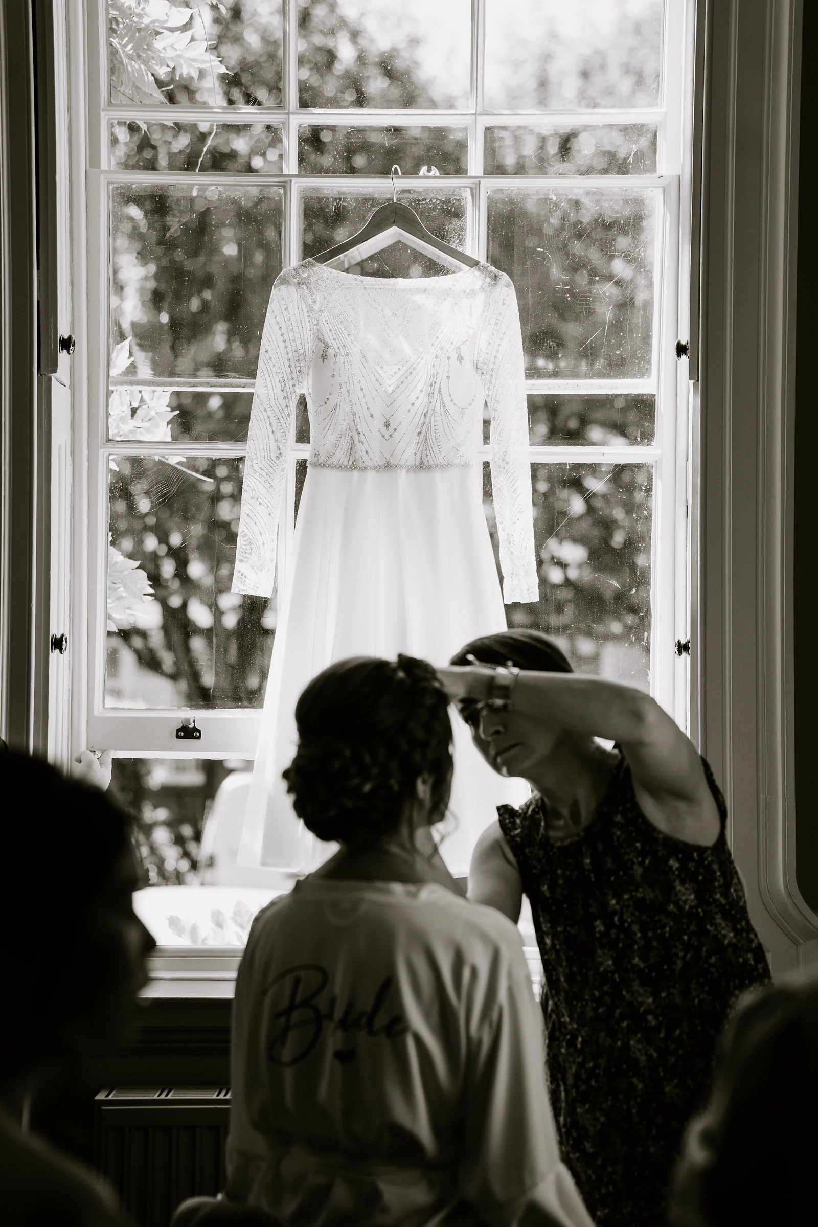 A wedding dress hangs at a tall window in black and white while the bride has her hair finished before the ceremony at The Orangery in Maidstone