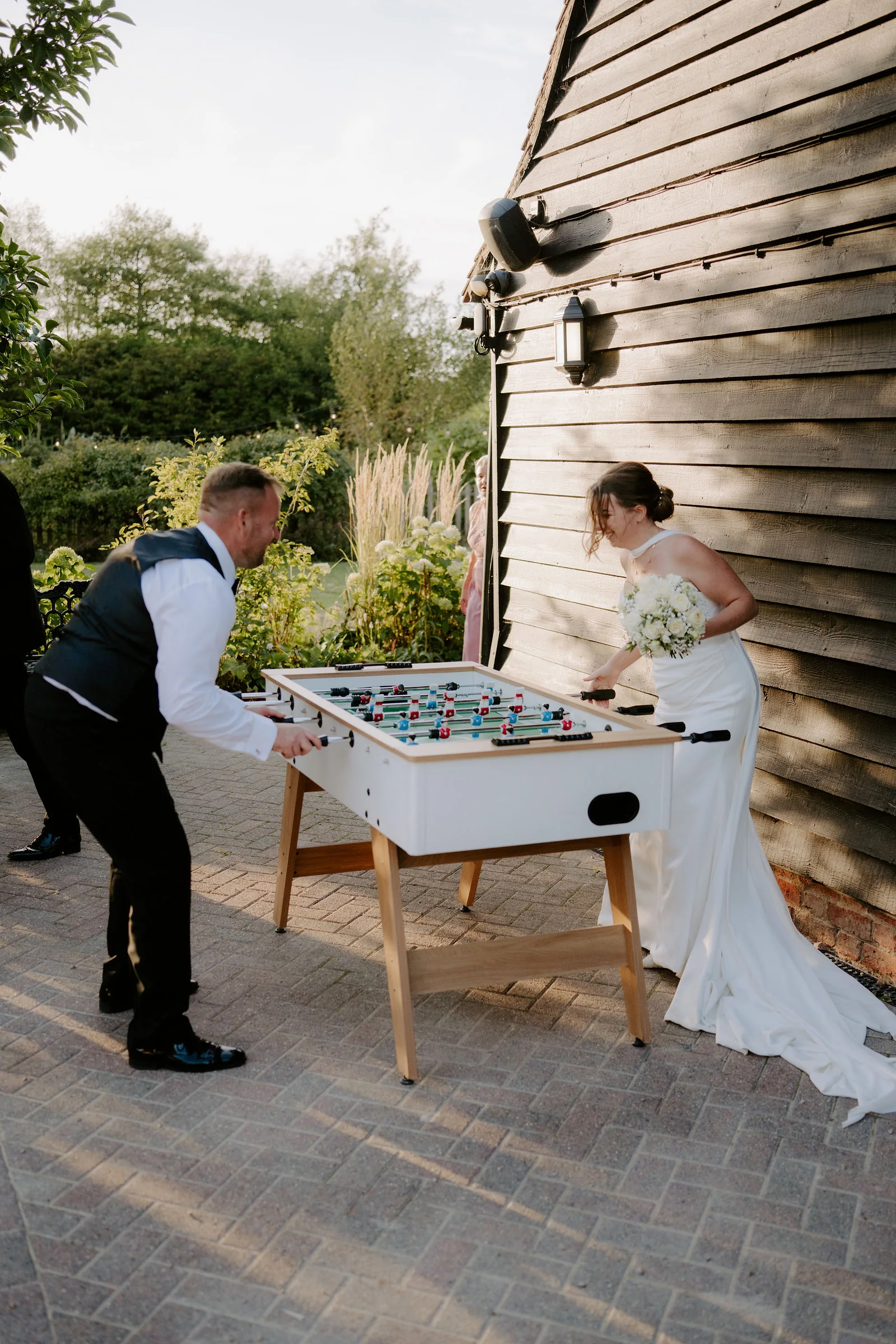 A groom in a waistcoat leans over a white table football table while his bride in a long gown plays opposite, on a paved yard beside a dark timber barn wall