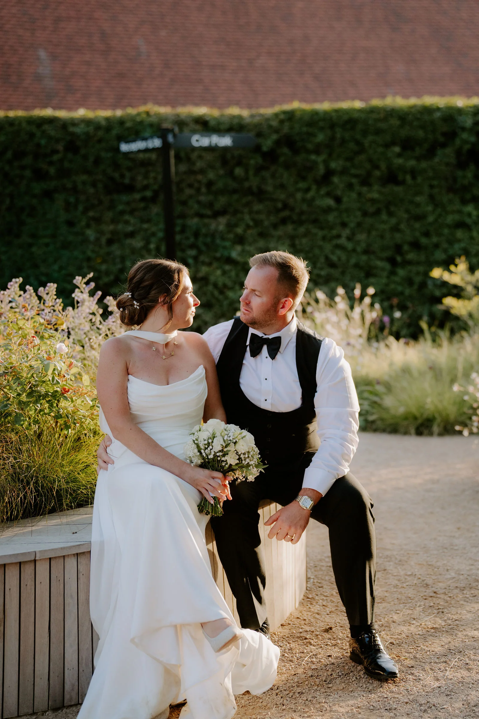 A bride in a strapless gown holding a bouquet sits on a low wooden bench beside her groom in a black waistcoat and bow tie, with a clipped hedge behind them in warm evening light
