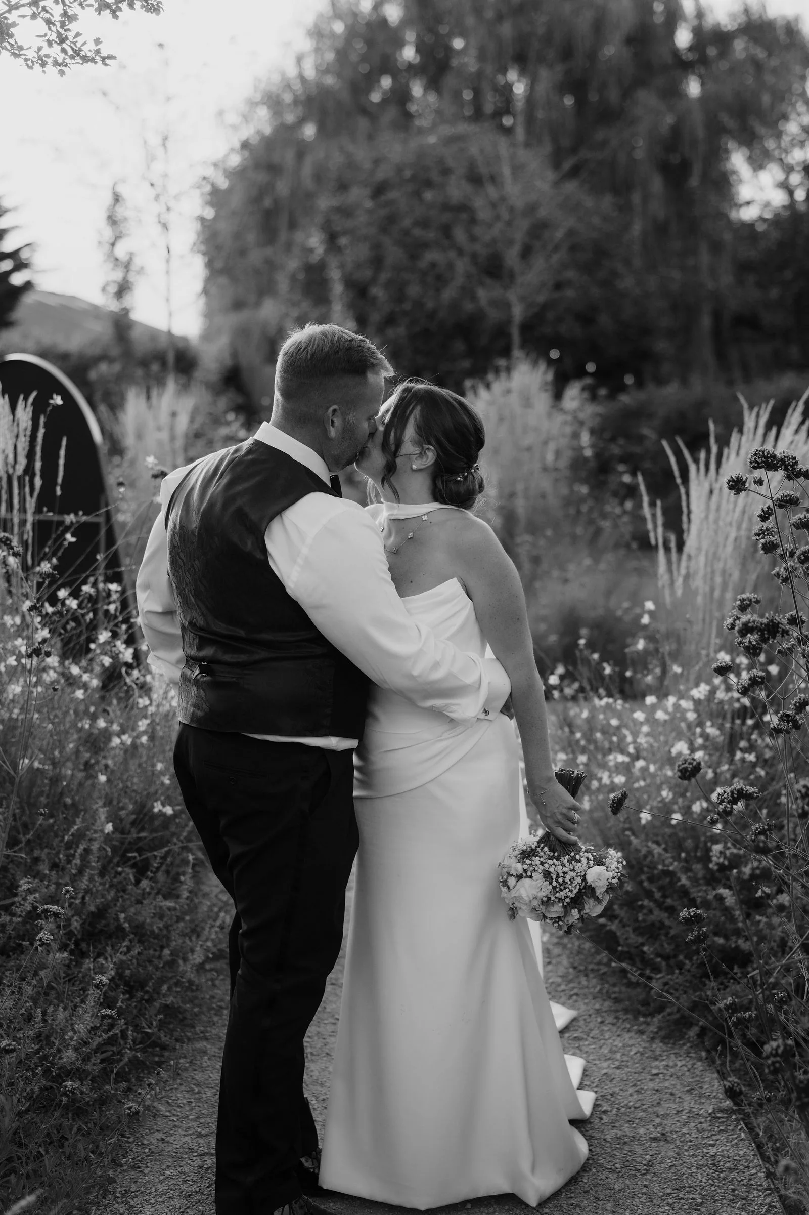 In black and white, a groom and a bride with a bouquet kiss on a gravel path between tall grasses and wildflowers