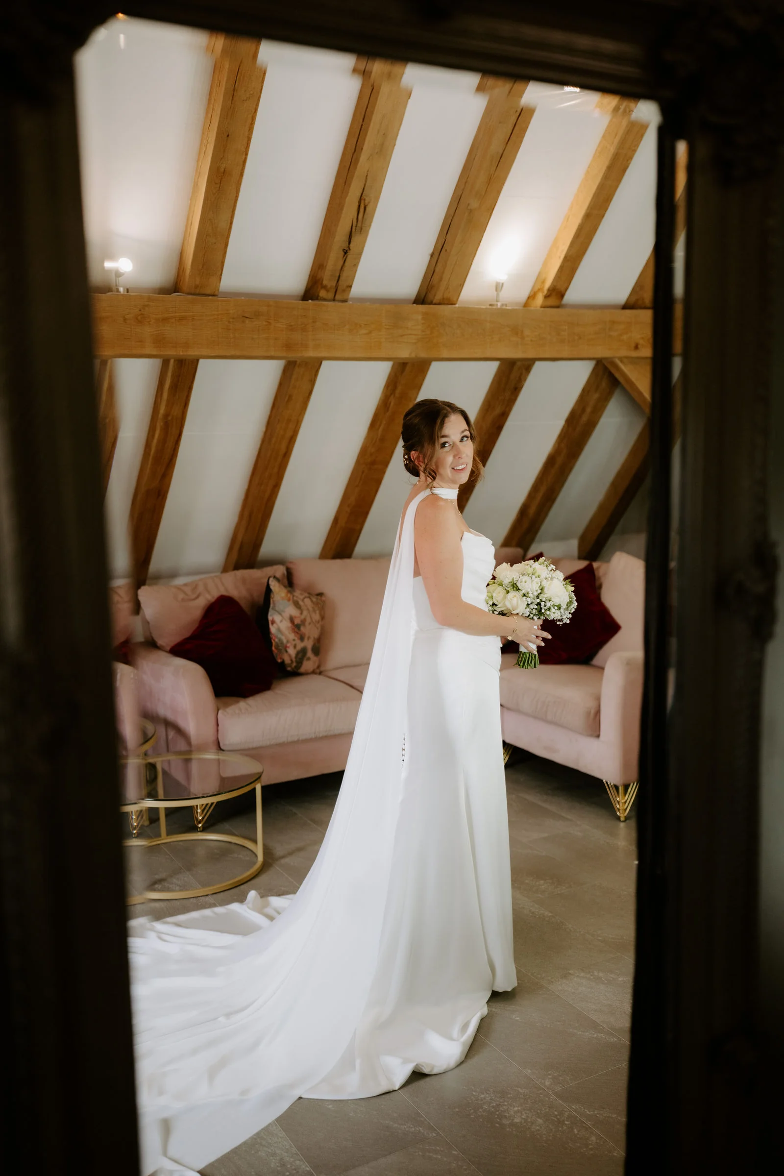 A bride in a fitted gown with a long cape holding a bouquet stands in a room with exposed oak roof beams and a pink sofa, seen through a dark frame
