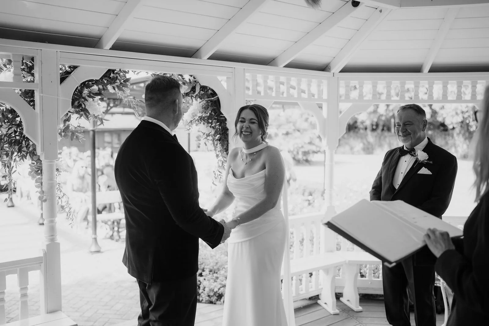 In black and white, a groom and a laughing bride hold hands facing each other inside a white garden gazebo as an officiant holds a book beside them
