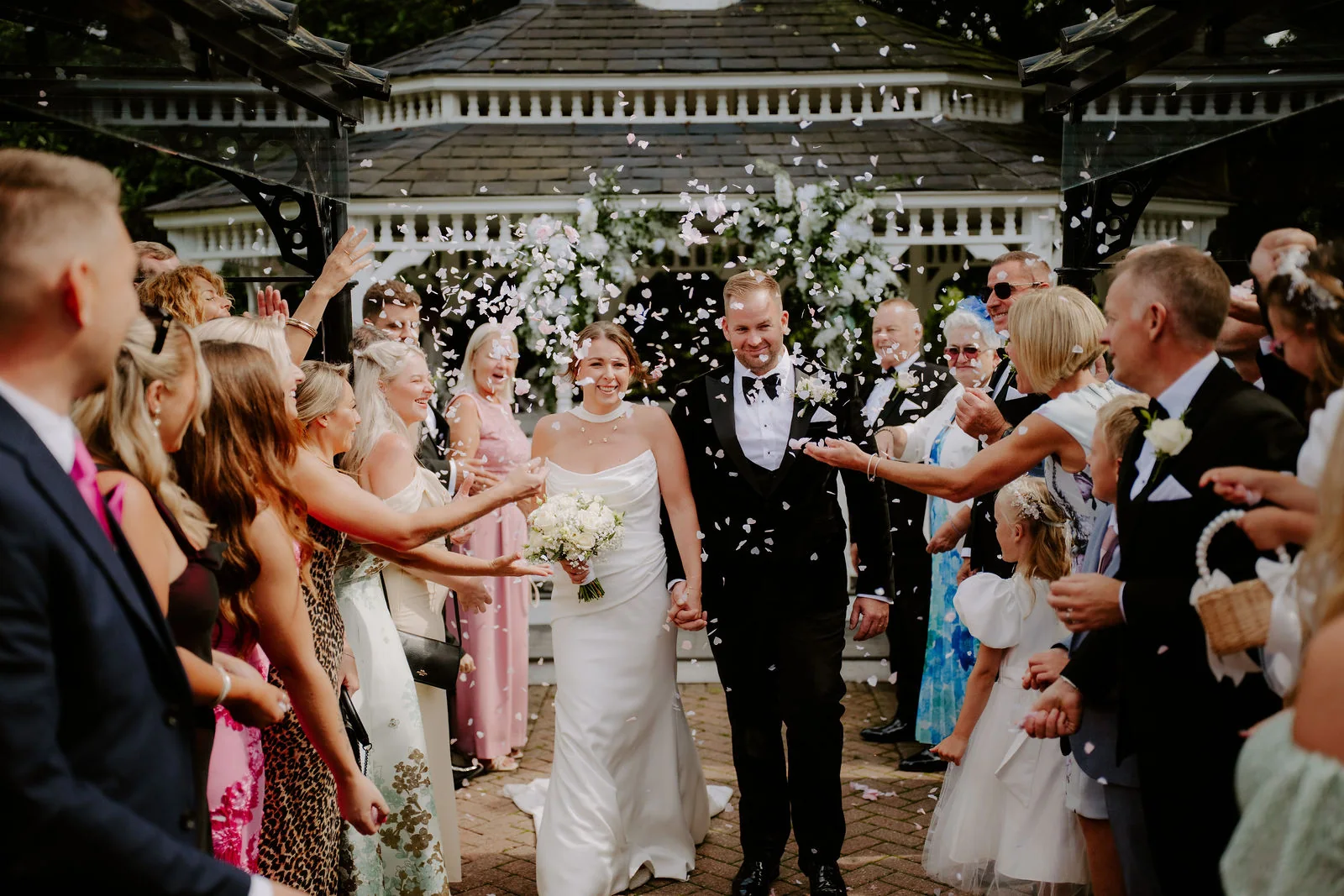 A bride in a strapless gown holding a bouquet and a groom in a black dinner suit walk out of a white garden gazebo between lines of guests throwing white petals