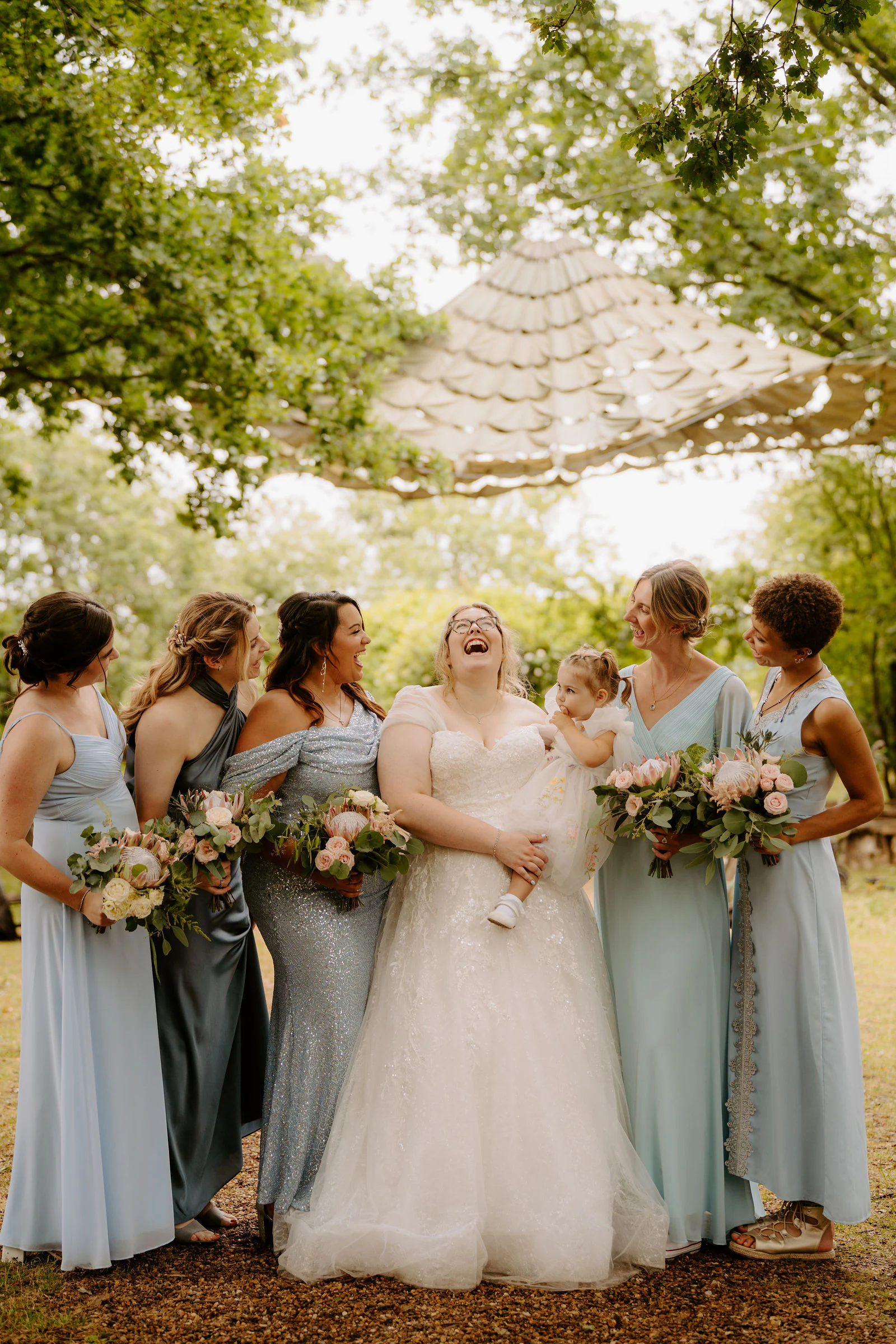 A bride laughs in the centre of her bridesmaids in pale blue dresses holding bouquets beneath trees at The Oak Grove near Sittingbourne