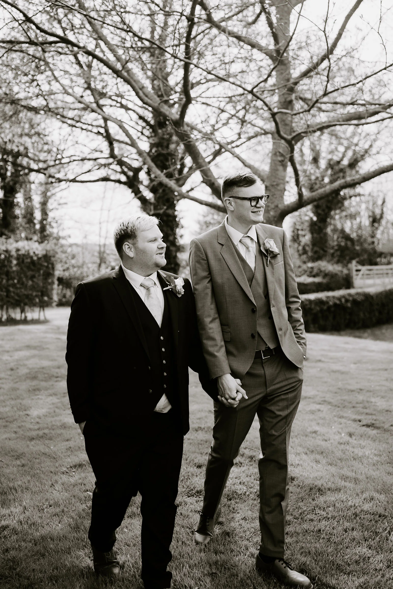 A black and white photograph of two grooms in three-piece suits walking hand in hand across a lawn beneath bare tree branches, both looking off to the side