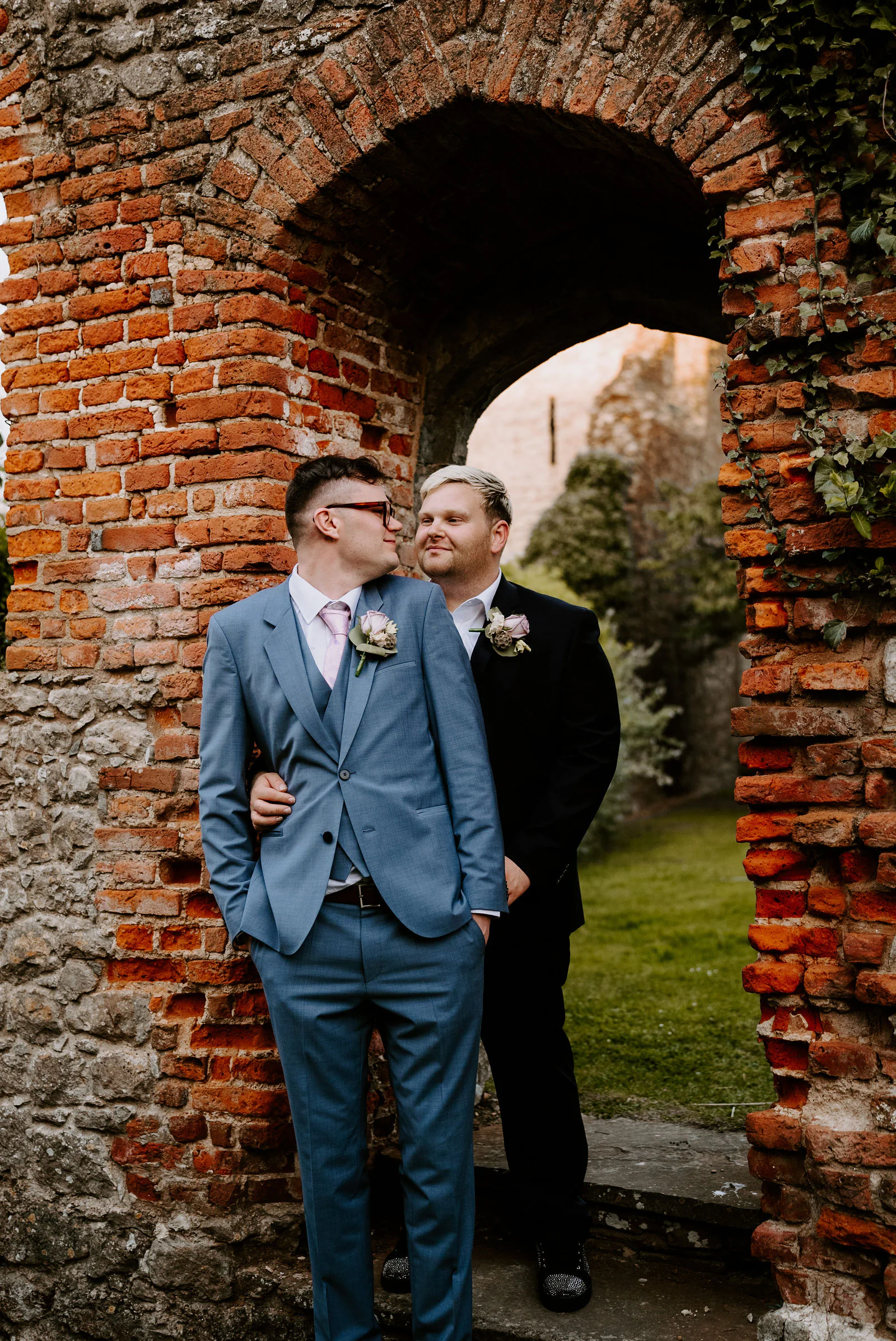 Two grooms stand close together in an ivy-covered old brick and flint archway at The Castle Westenhanger, one in a light blue suit and glasses and the other in a black suit, looking at each other