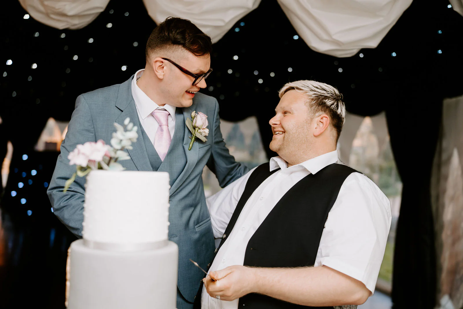 Two grooms laugh as they cut a white two-tier wedding cake topped with pale flowers beneath a draped ceiling of fairy lights