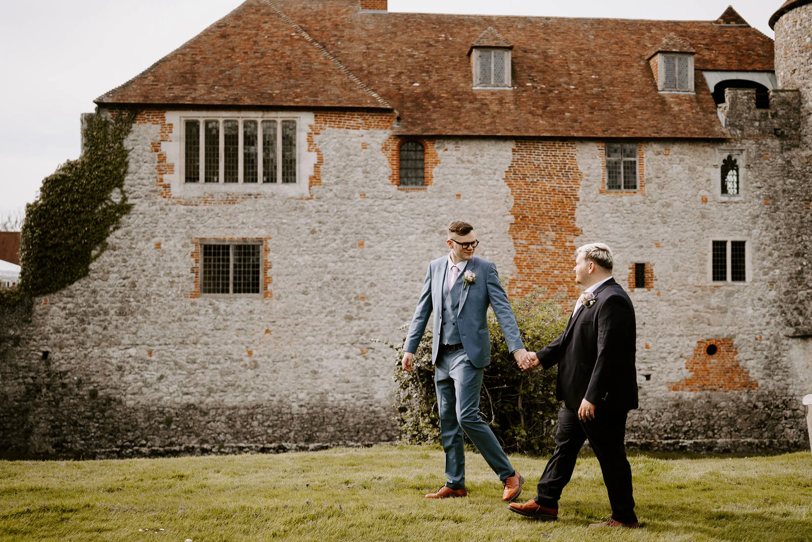 Two grooms hold hands and walk across a lawn, looking at each other, in front of an old flint and brick building with a tiled roof and a stone turret at The Castle Westenhanger