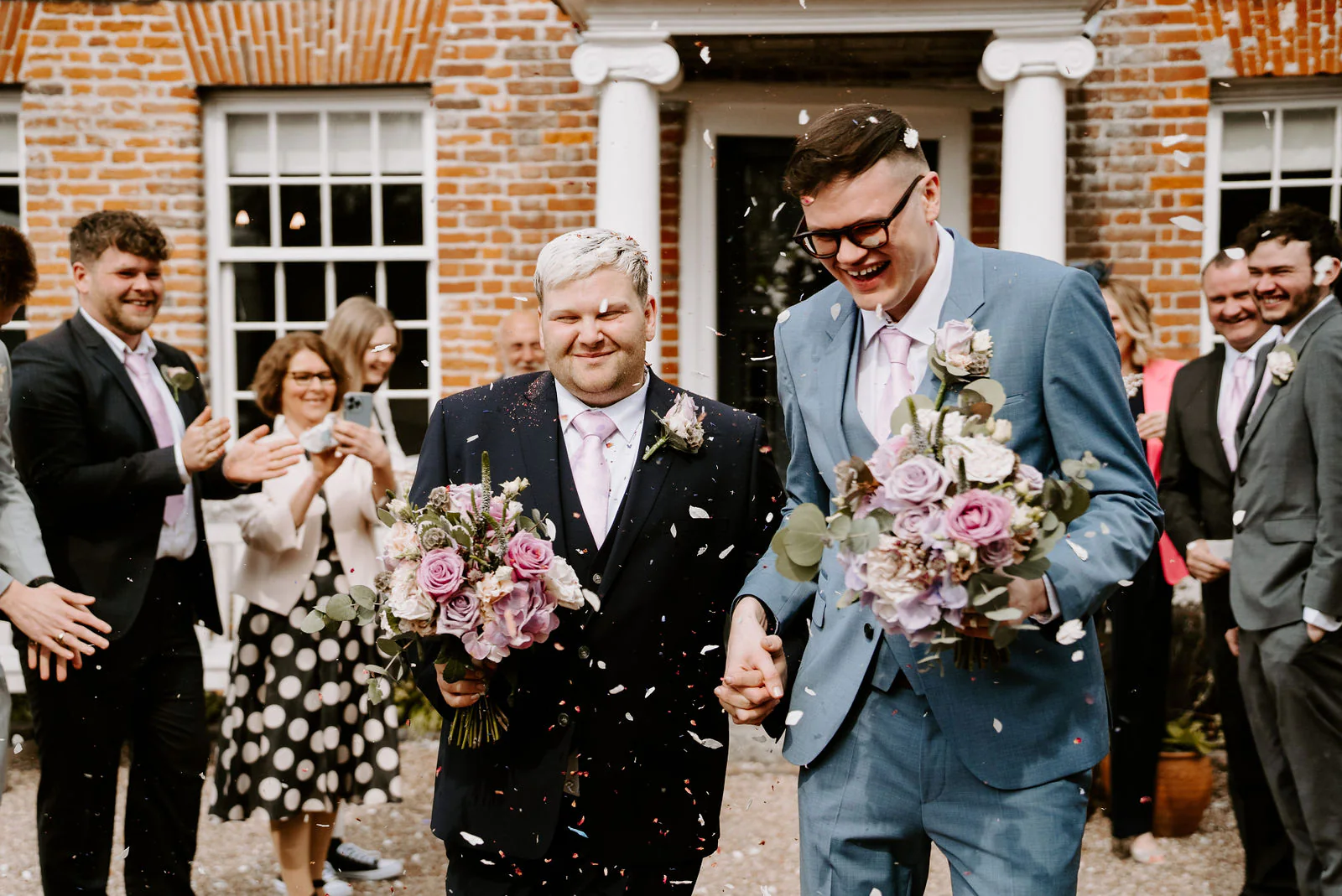 Two grooms hold hands and laugh as confetti falls around them outside a red brick building with a white pillared doorway, each holding a bouquet of pink and lilac flowers, with guests clapping beside them