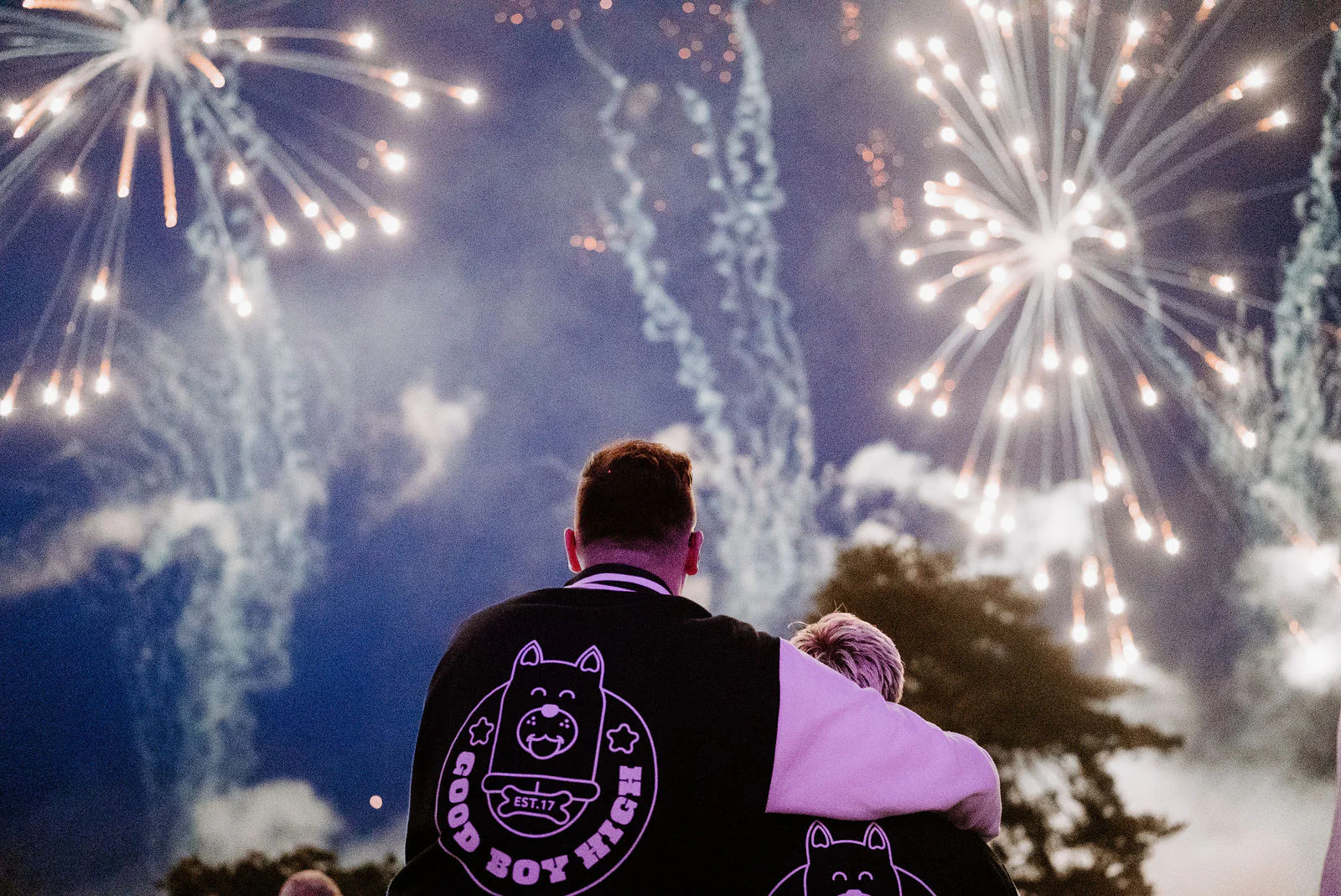 Seen from behind, two grooms in matching black jackets with lilac sleeves stand with their arms around each other and look up at fireworks bursting over the trees at night