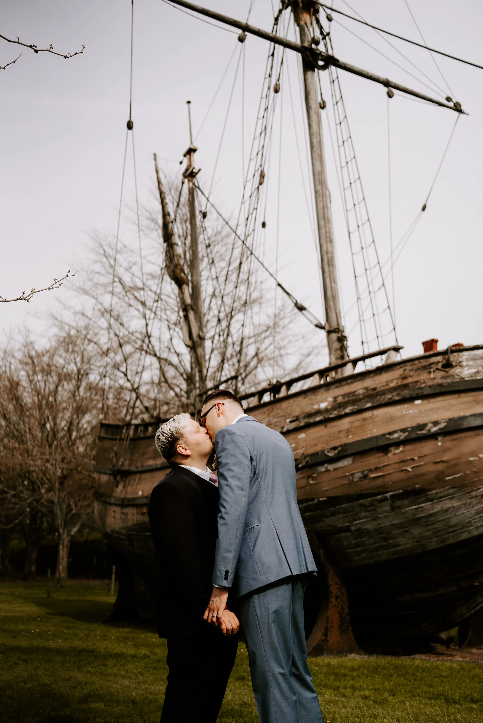 Two grooms kiss on a lawn in front of a weathered wooden sailing ship resting on dry land, with its masts and rigging rising behind them